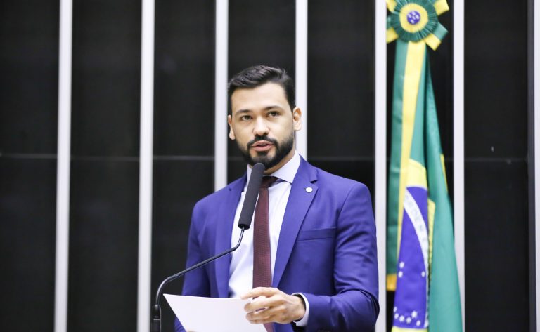 Deputado Samuel Viana fala ao microfone. Ele é um homem claro com cabelo e barba pretos, usa um terno azul escuro e ao fundo no lado direito há uma bandeira do Brasil