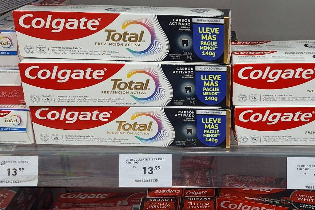 A imagem mostra prateleiras de produtos de higiene bucal, especificamente tubos de creme dental da marca Colgate. Os tubos estão organizados em duas fileiras, com destaque para o produto