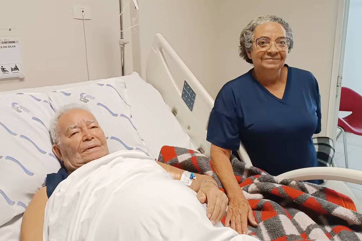 A imagem mostra um homem idoso deitado em uma cama de hospital, coberto com um lençol branco e um cobertor xadrez vermelho e preto. Ao lado dele, uma mulher idosa está em pé, sorrindo. Ambos parecem estar em um ambiente hospitalar, com paredes claras e equipamentos médicos ao fundo.