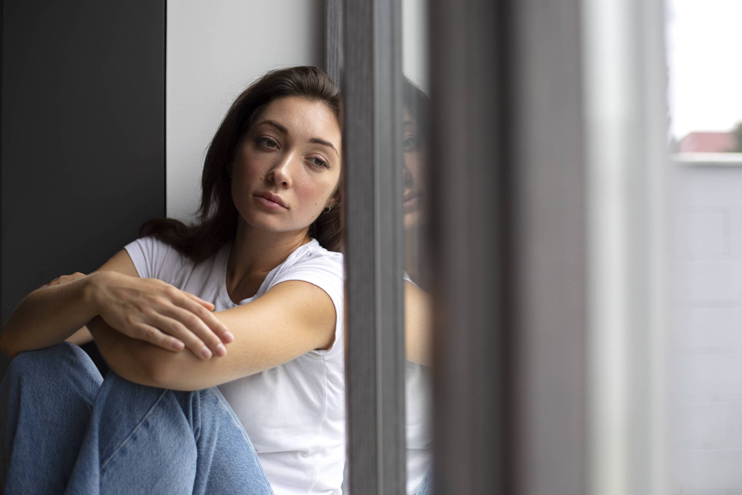 Uma mulher está sentada encostada em uma parede, com as pernas dobradas e os braços envolvendo os joelhos. Ela tem cabelo castanho escuro e está usando uma camiseta branca. A expressão dela é pensativa e um pouco melancólica. Ao fundo, há uma janela com luz natural entrando, refletindo parte do ambiente.