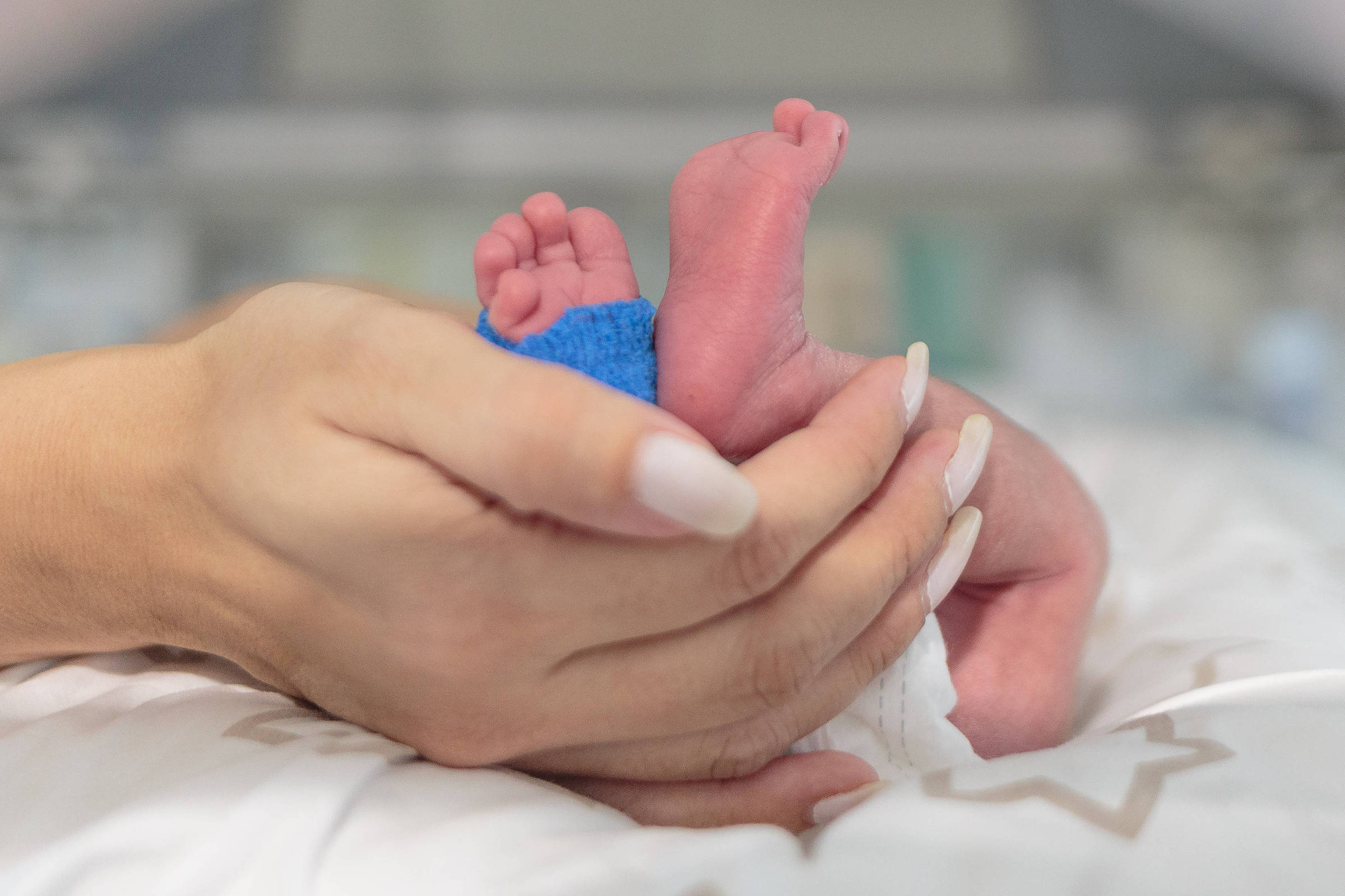A imagem mostra uma mão humana segurando o pé de um bebê. O pé do bebê está parcialmente coberto por uma pequena peça de roupa azul. O fundo é desfocado, mas parece ser um ambiente hospitalar, possivelmente uma unidade de terapia intensiva neonatal. A mão que segura o pé é feminina, com unhas bem cuidadas e pintadas.