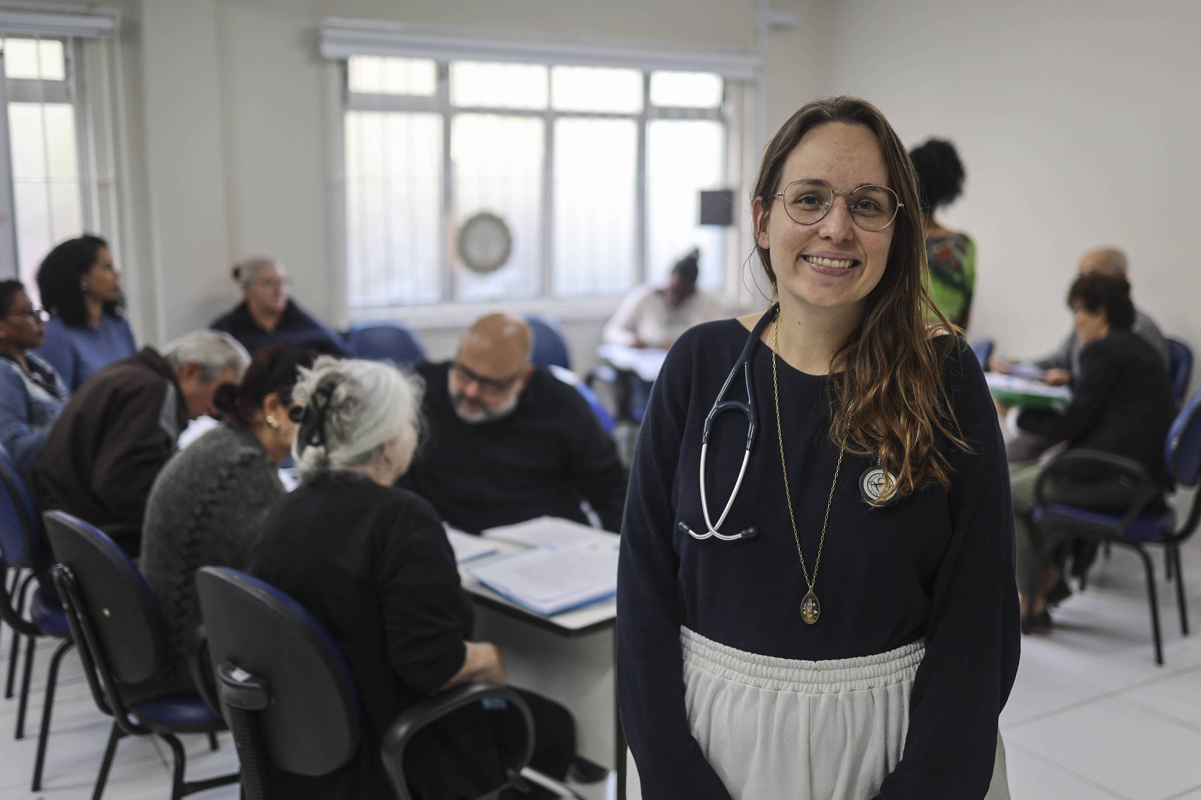 Uma mulher com óculos e um estetoscópio ao redor do pescoço sorri para a câmera. Ela está em um ambiente de sala de aula ou reunião, onde várias pessoas estão sentadas em cadeiras, algumas delas voltadas para uma mesa com documentos. A sala tem janelas grandes que permitem a entrada de luz natural, e as pessoas parecem estar envolvidas em uma discussão ou atividade em grupo.
