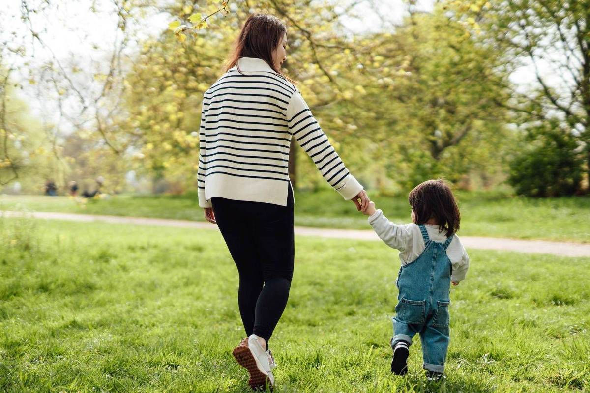 Uma mulher e uma criança estão caminhando de mãos dadas em um parque. A mulher usa um suéter listrado e calças pretas, enquanto a criança está vestida com um macacão azul e uma blusa branca. O cenário é verde, com árvores ao fundo e um caminho visível.