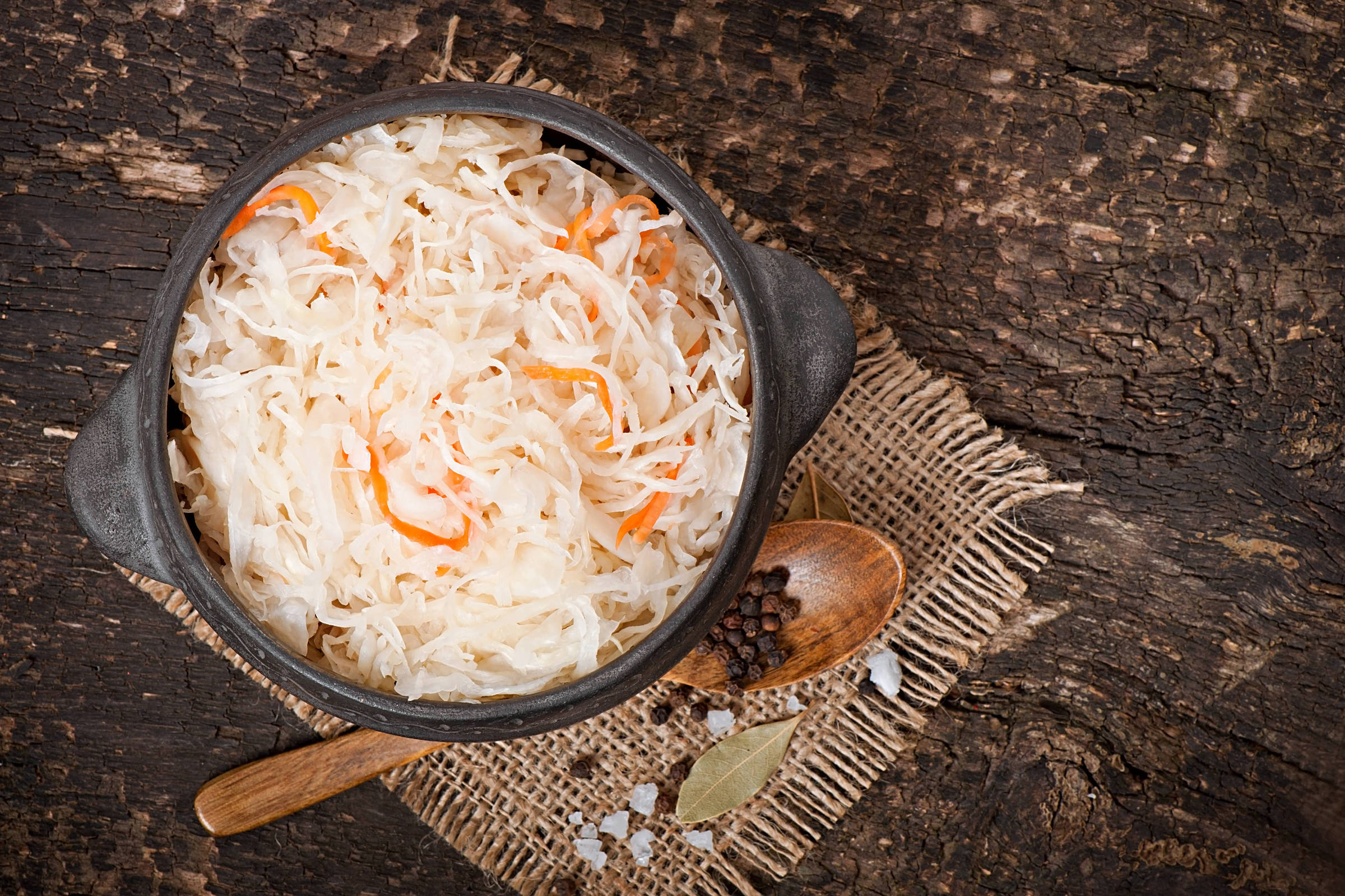 Uma panela de barro com chucrute, que é uma mistura de repolho fermentado e cenoura. A panela está sobre uma superfície de madeira escura, e ao lado há uma colher de madeira com grãos de pimenta e uma folha de louro. O fundo é texturizado com um pano de juta.