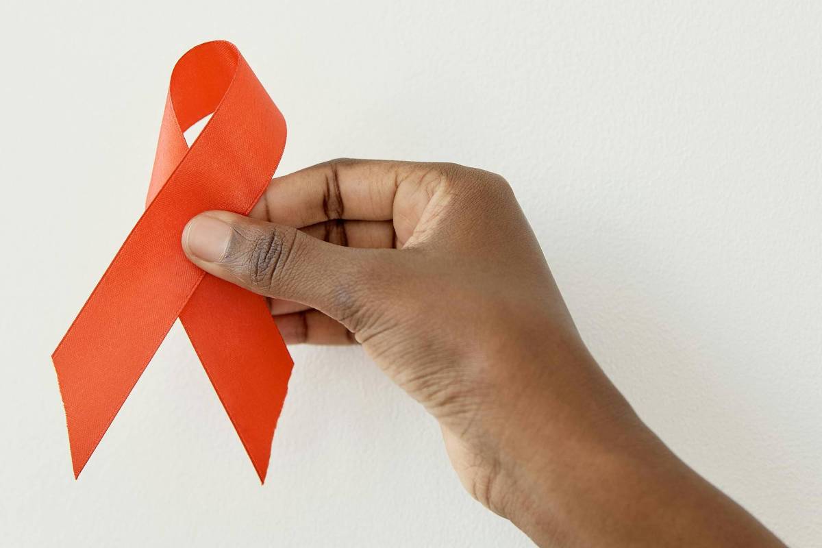 Uma mão segurando um laço vermelho em frente a um fundo branco. O laço é feito de fita e está em uma posição vertical, simbolizando a conscientização sobre a AIDS.