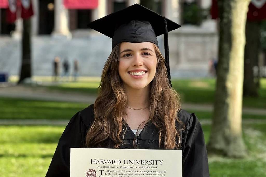 Uma jovem mulher está sorrindo enquanto segura um diploma da Harvard University. Ela está vestida com uma toga de formatura e um capelo preto. Ao fundo, há bandeiras da universidade e árvores, indicando um ambiente de campus universitário. A iluminação é natural, sugerindo que a foto foi tirada durante o dia.