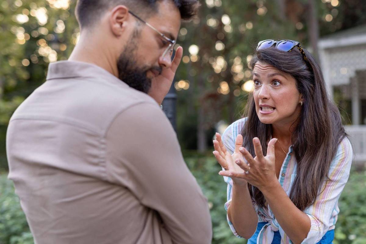 A imagem mostra um homem e uma mulher em um ambiente externo, possivelmente em um parque. A mulher está gesticulando com as mãos e parece estar expressando emoções intensas, enquanto o homem está com a cabeça baixa, cobrindo o rosto com uma das mãos, indicando que está pensativo ou preocupado. Ao fundo, há uma estrutura que parece ser uma casa ou gazebo, cercada por vegetação.
