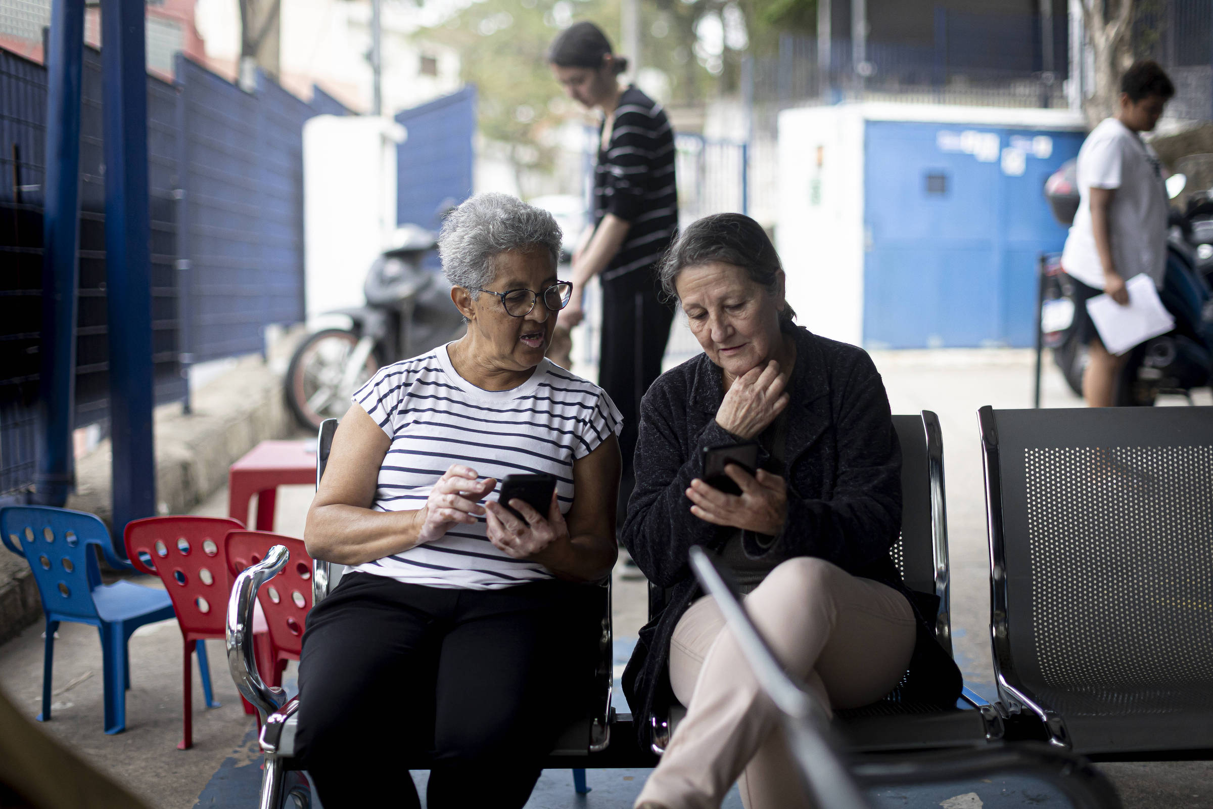 Duas mulheres sentadas em cadeiras, uma ao lado da outra, olhando para seus celulares. A mulher à esquerda tem cabelo grisalho e usa uma blusa listrada, enquanto a mulher à direita tem cabelo escuro e usa uma blusa preta. Ao fundo, há uma parede azul e algumas motos estacionadas. O ambiente parece ser um espaço público, com algumas cadeiras vermelhas e azuis visíveis.
