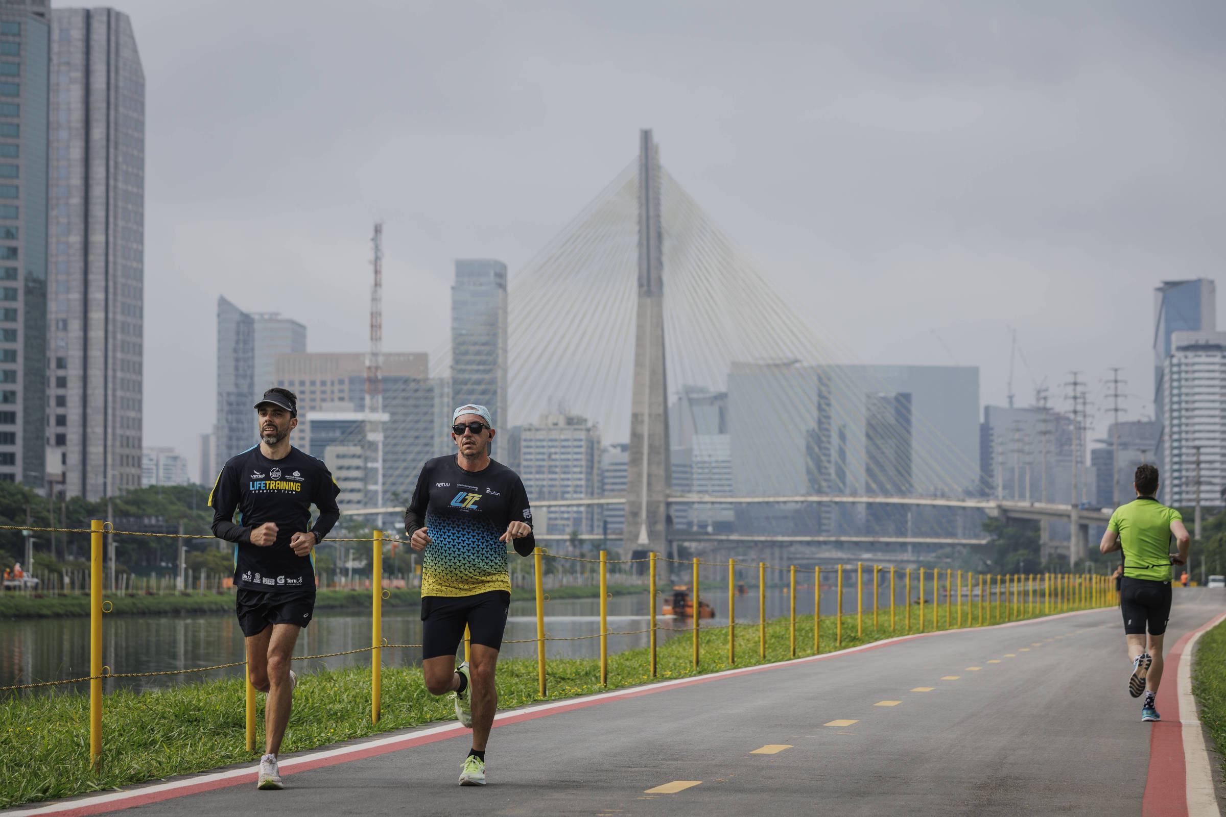 A imagem mostra dois homens correndo em uma pista ao lado de um rio, com a cidade de São Paulo ao fundo. O primeiro corredor está usando uma camiseta preta e um boné, enquanto o segundo usa uma camiseta preta com detalhes em amarelo e um óculos de sol. Ao fundo, é possível ver a Ponte Estaiada e vários edifícios altos, característicos da paisagem urbana de São Paulo. O clima parece nublado e a pista é pavimentada com uma faixa vermelha.