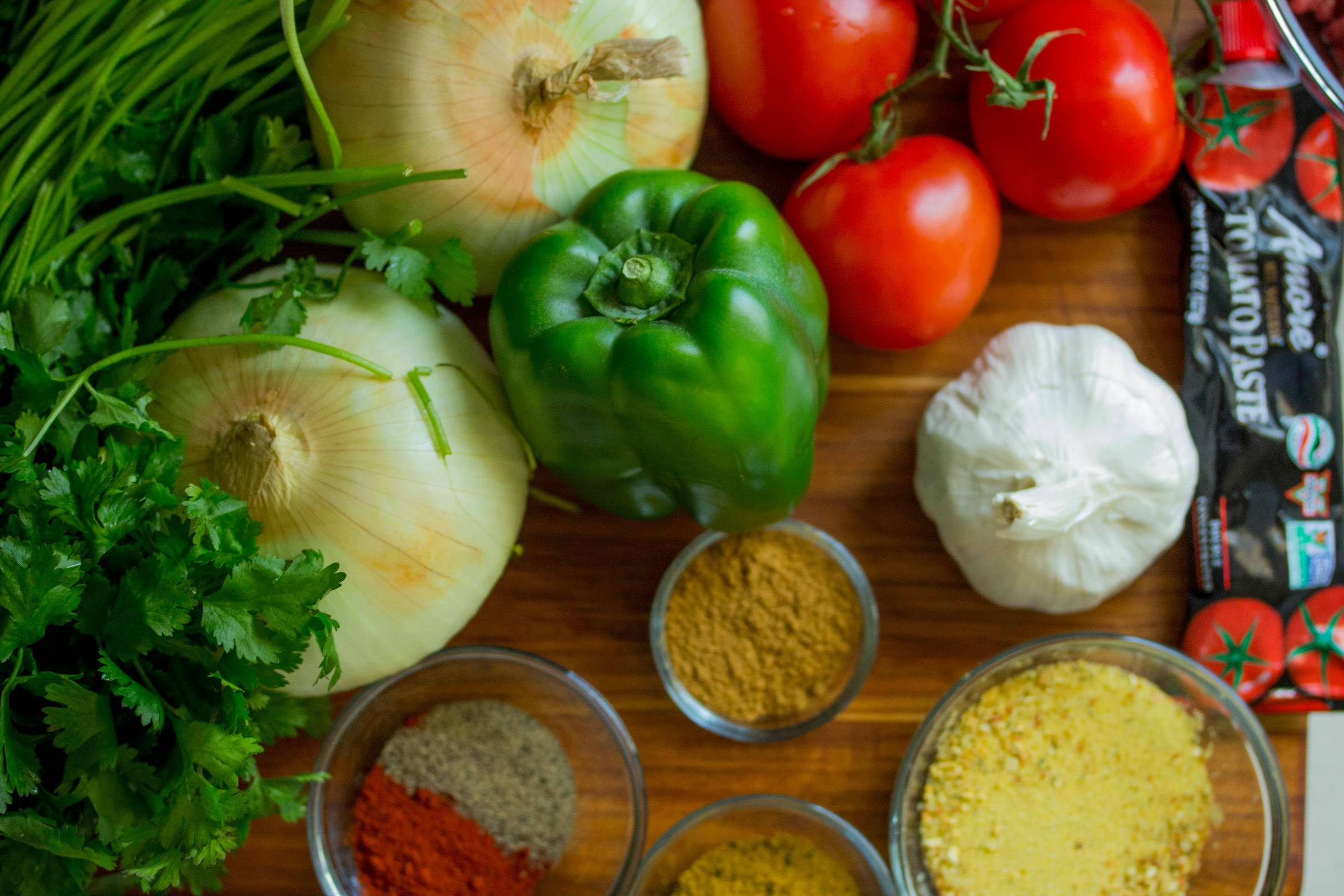 A imagem mostra uma variedade de ingredientes frescos dispostos sobre uma superfície de madeira. Há cebolas brancas, pimentão verde, tomates vermelhos, um bulbo de alho e um punhado de coentro. Também estão presentes várias tigelas com especiarias em pó, incluindo curry, cominho, pimenta e outros temperos.