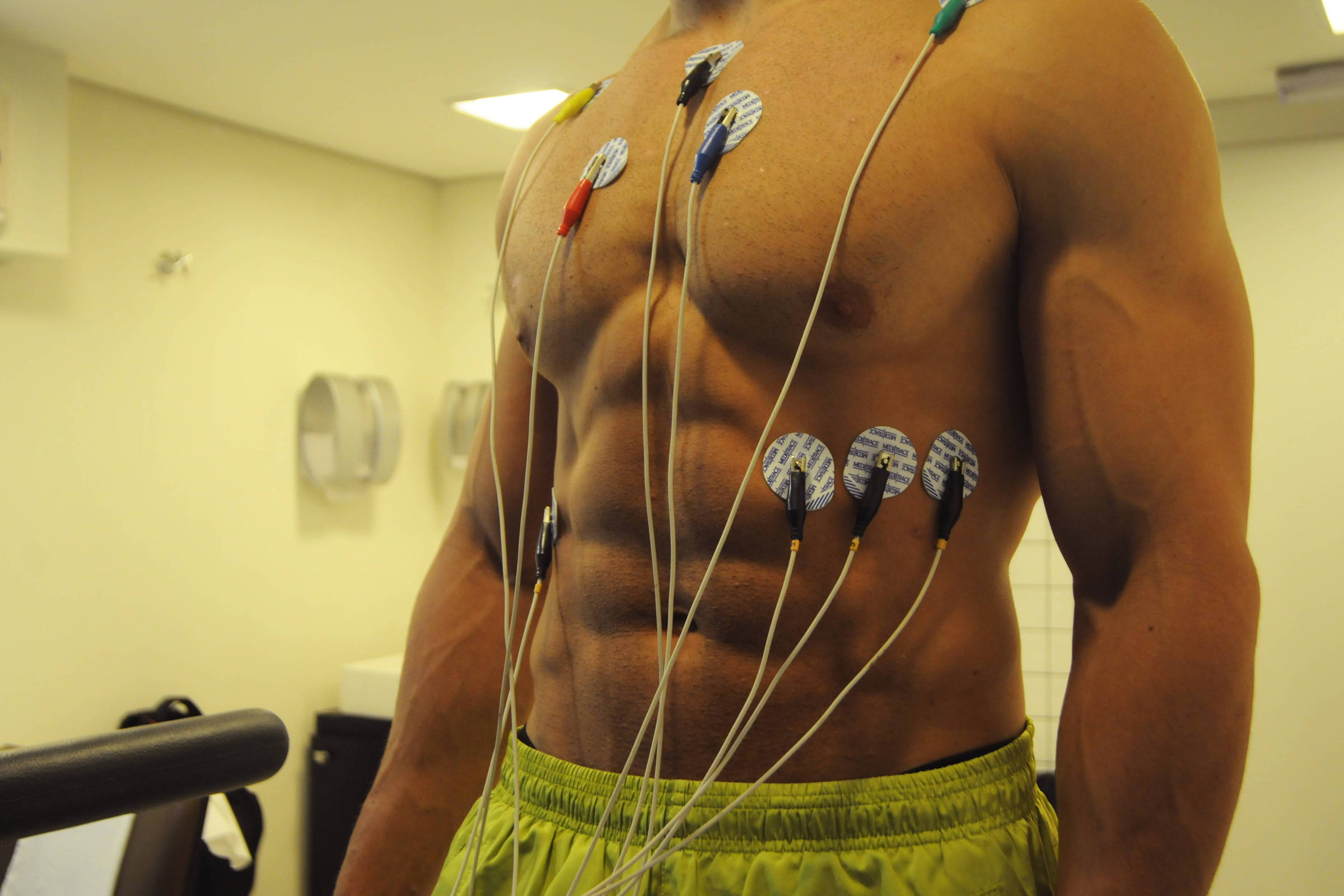 A imagem mostra um homem com um corpo musculoso, em pé, com eletrodos adesivos colocados em seu peito e abdômen. Os eletrodos estão conectados a fios que se estendem para fora da imagem. O ambiente é uma sala de testes, com paredes claras e alguns equipamentos visíveis ao fundo. O homem está usando um short amarelo.