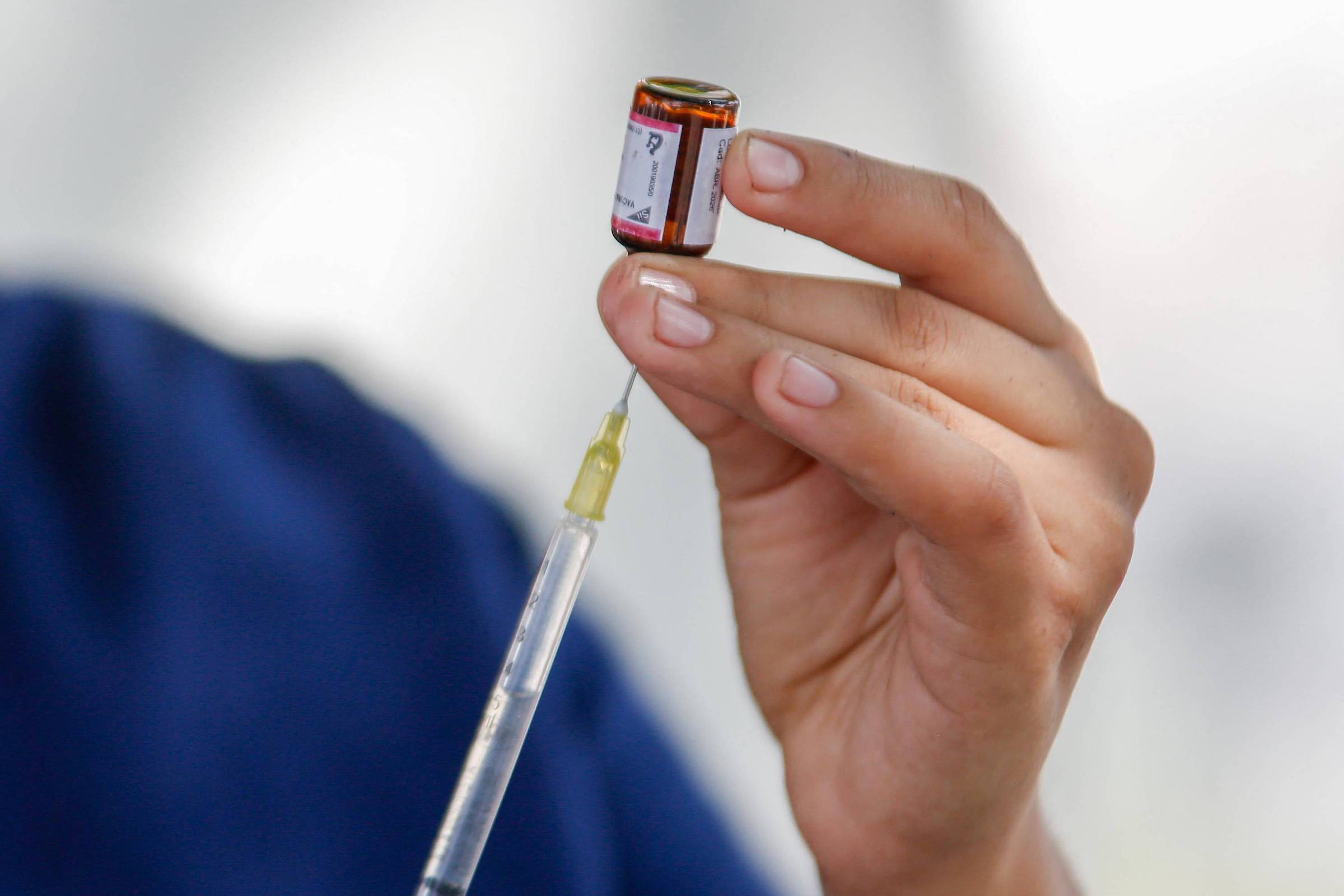 Uma profissional de saúde está segurando uma seringa e um frasco de vacina. A seringa está parcialmente preenchida com o líquido da vacina, enquanto a profissional observa atentamente o frasco. O fundo é desfocado, sugerindo um ambiente de vacinação.