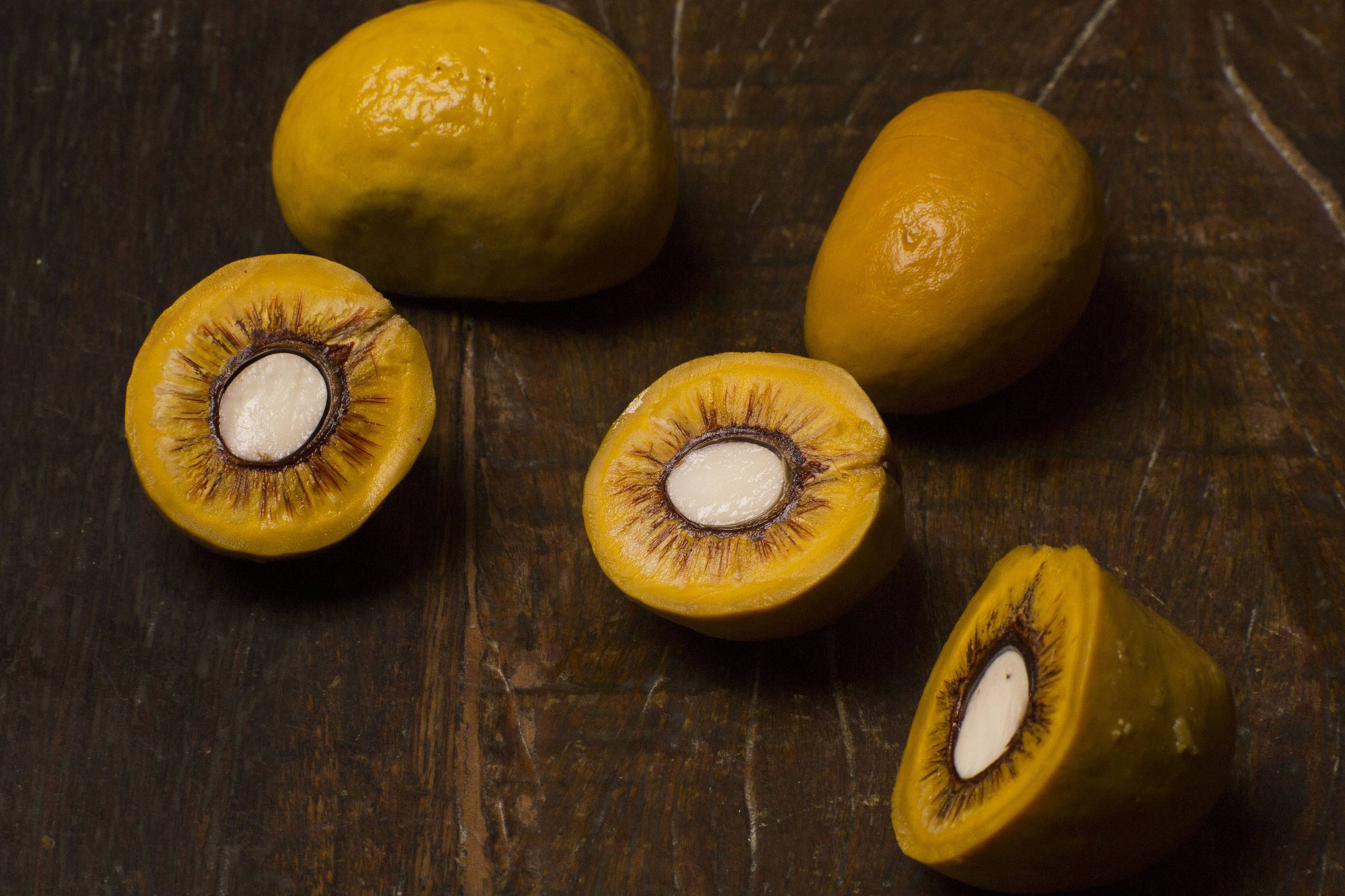 A imagem mostra várias frutas amarelas, algumas inteiras e outras cortadas ao meio. As frutas cortadas revelam uma polpa amarela com uma semente branca no centro, cercada por uma casca fina e marrom. O fundo é uma superfície de madeira escura.