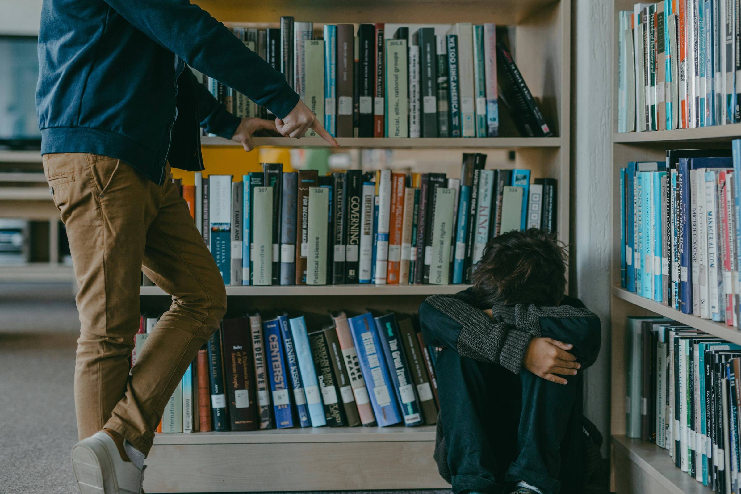 A imagem mostra uma cena em uma biblioteca. À esquerda, uma pessoa em pé, vestindo uma jaqueta azul e calças claras, está apontando para uma estante de livros. À direita, uma pessoa está sentada no chão, encostada na estante, com os joelhos dobrados e os braços cobrindo a cabeça, aparentando estar em uma posição de defesa ou tristeza. A estante está cheia de livros organizados, e o ambiente parece calmo, mas a interação entre as duas pessoas sugere uma situação de bullying.