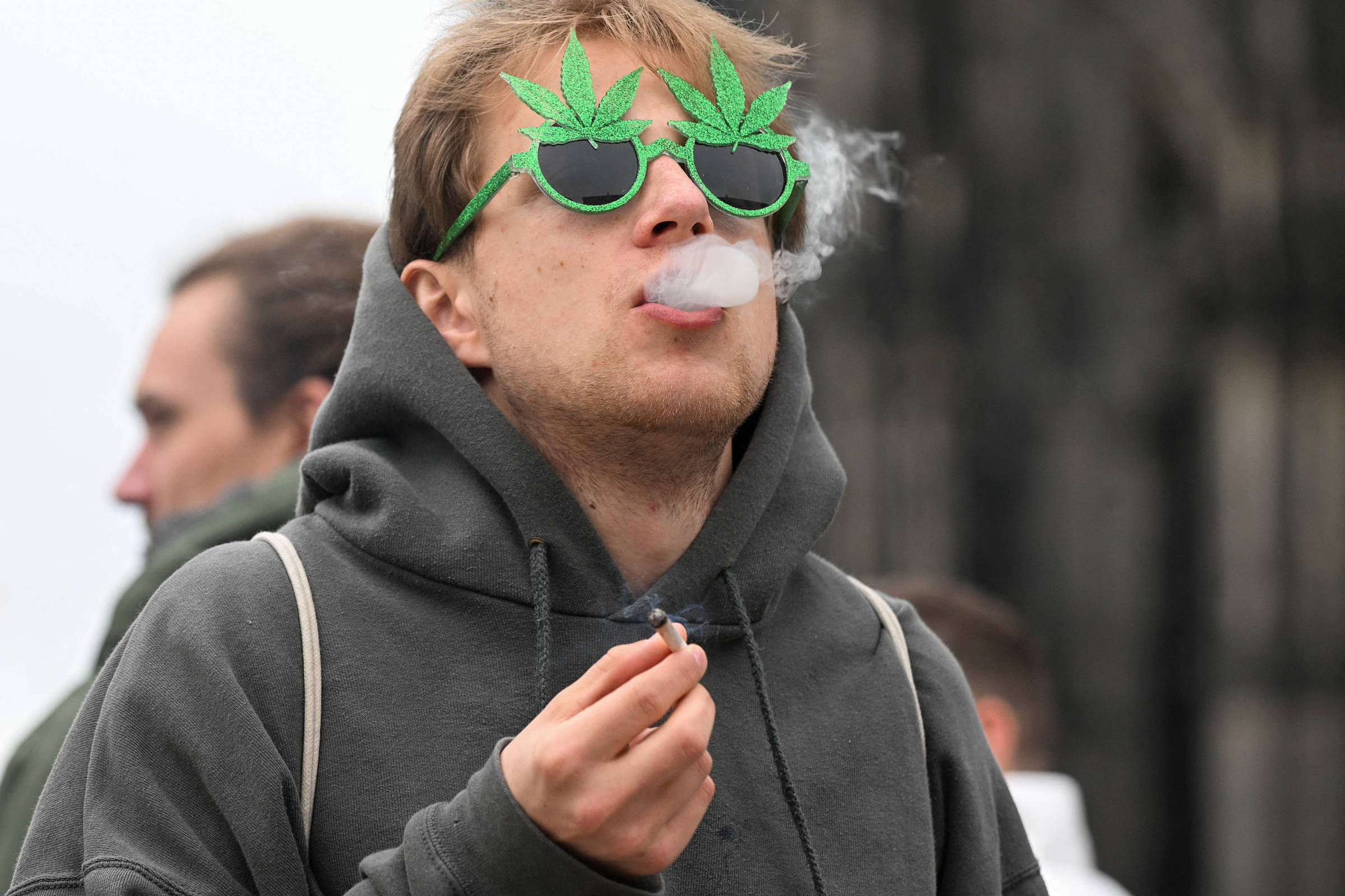Um jovem está fumando e exalando fumaça. Ele usa um moletom cinza e óculos de sol decorados com folhas de maconha. Ao fundo, outras pessoas estão visíveis, mas não são o foco da imagem.