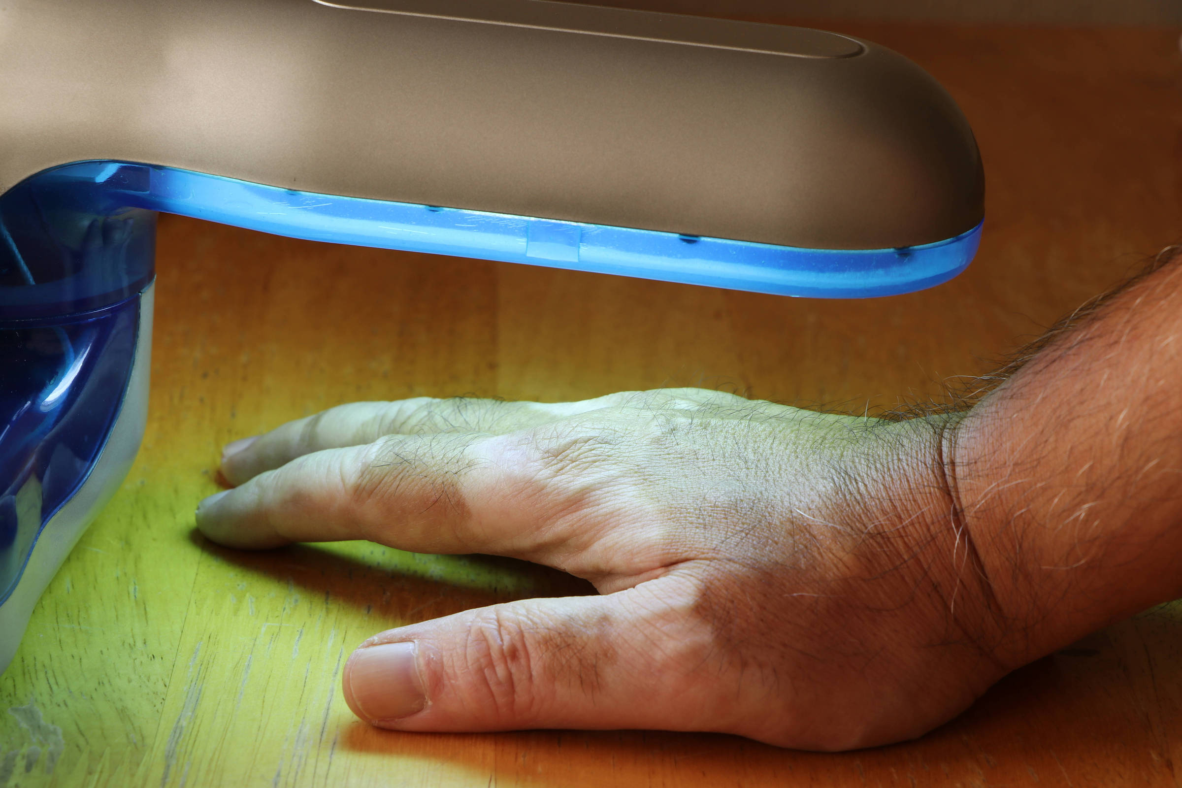A imagem mostra uma mão humana colocada sobre uma superfície de madeira, sob uma lâmpada UV que emite uma luz azul. A lâmpada está posicionada acima da mão, que está relaxada e com a palma voltada para baixo. A base da lâmpada é de cor azul e prata, e a luz azul ilumina a área ao redor da mão.