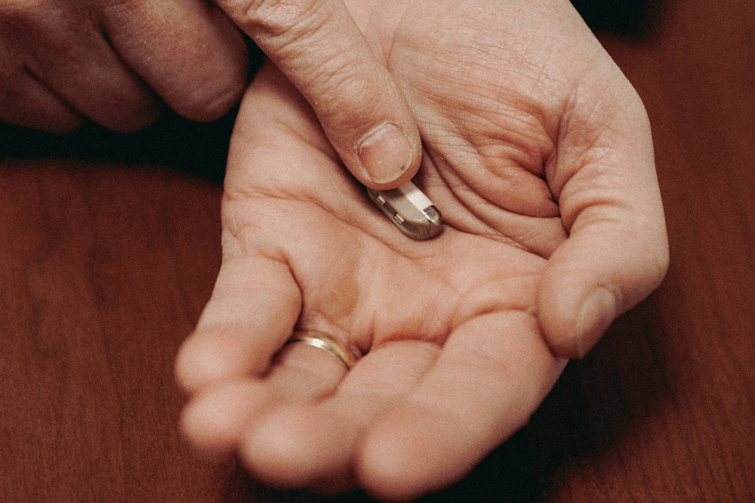 Uma mão masculina, com um anel no dedo, está segurando um objeto prateado na palma. Outra mão está apontando para o objeto. O fundo é uma superfície de madeira.