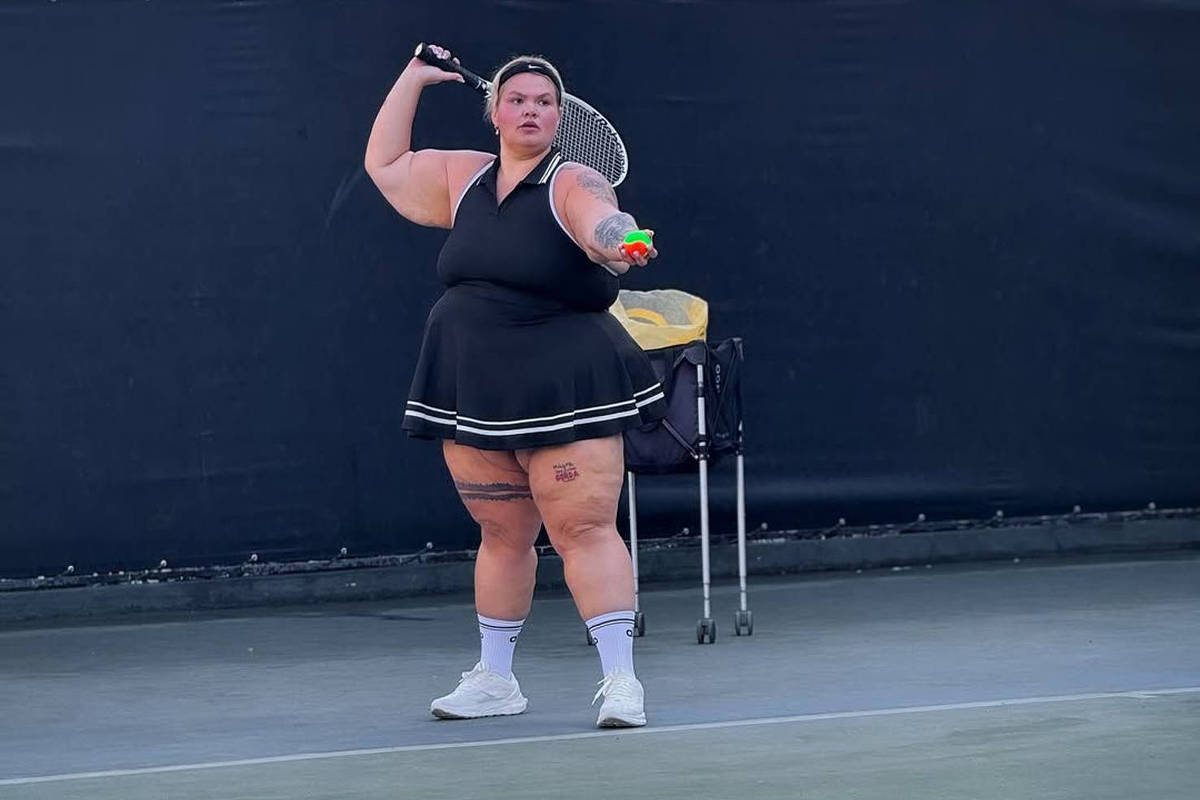 Uma mulher está jogando tênis em uma quadra. Ela usa um vestido preto com detalhes brancos e está segurando uma raquete. A mulher parece estar se preparando para fazer um saque, com uma expressão concentrada. Ao fundo, há uma rede de tênis e um cenário urbano visível, com prédios ao longe. A quadra é de superfície verde e há um carrinho de equipamentos ao lado.