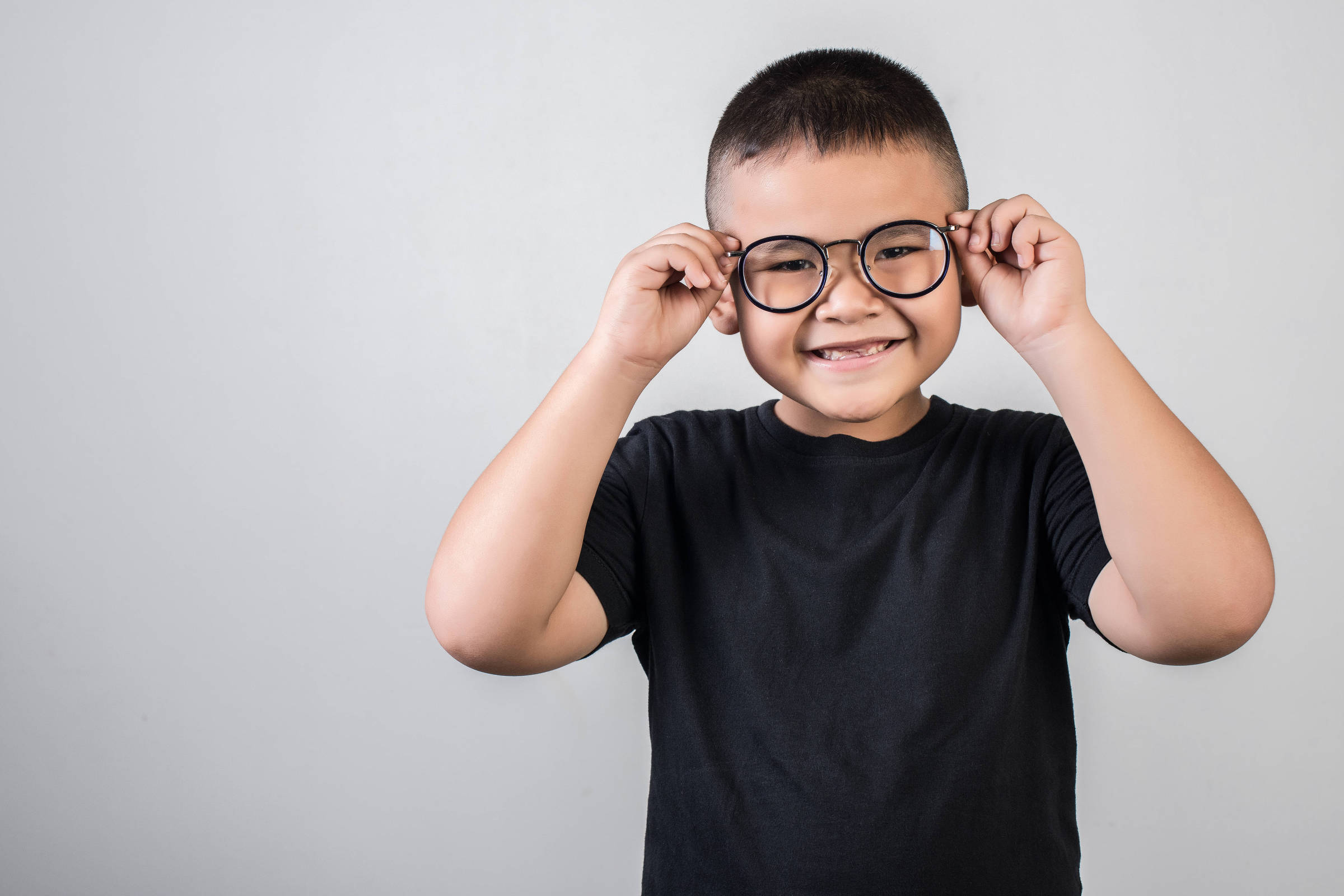 Um menino sorridente está posando para a foto, segurando seus óculos com as duas mãos. Ele usa uma camiseta preta e tem cabelo curto. O fundo da imagem é cinza claro, criando um contraste com a roupa do menino.