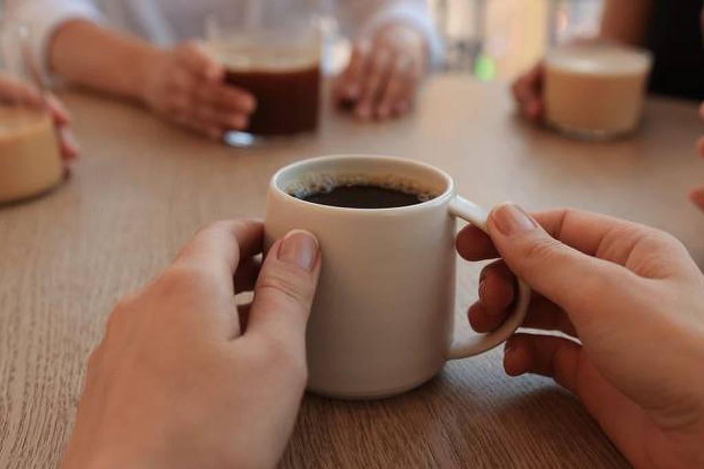 A imagem mostra uma mesa de madeira com várias mãos segurando copos de café e bebidas. Em primeiro plano, uma mão segura uma caneca branca com café preto. Ao fundo, outras pessoas estão segurando copos de diferentes tipos de bebidas, como café com leite e outras variações. A atmosfera parece ser de um encontro social ou uma conversa descontraída.