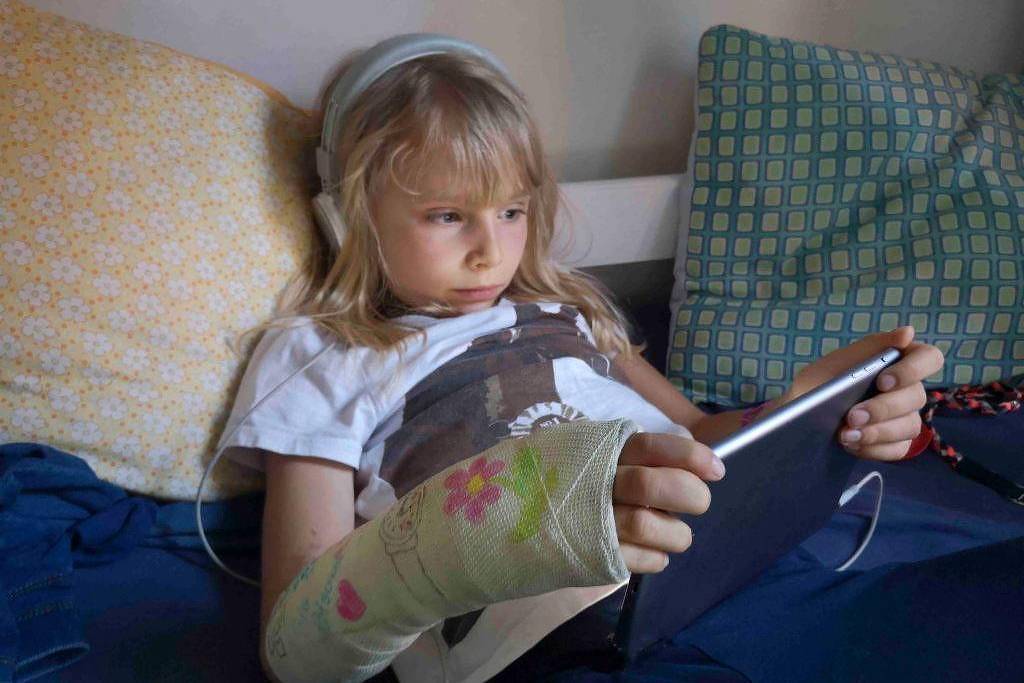 Uma criança está sentada em uma cama, usando um tablet. Ela tem fones de ouvido e uma camisa branca. O braço esquerdo está imobilizado com uma atadura colorida. Ao fundo, há duas almofadas, uma amarela e outra com padrão quadriculado.