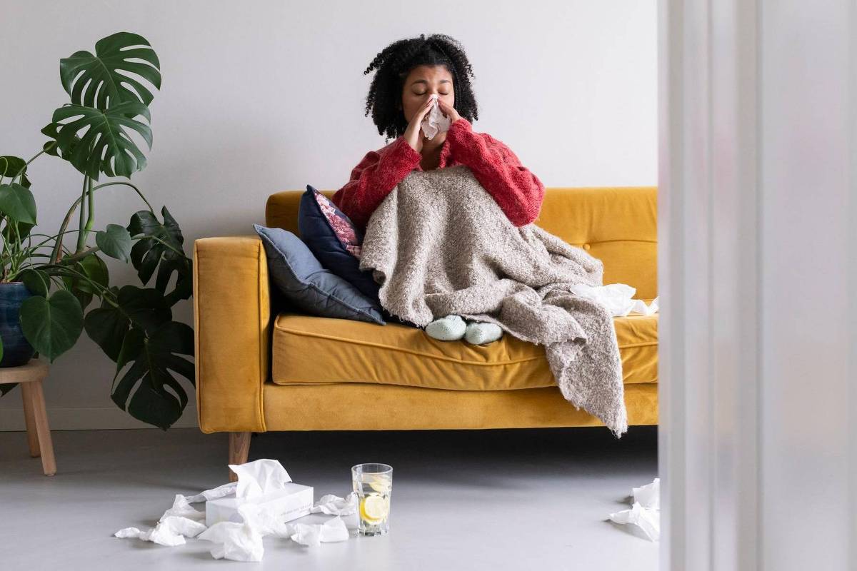 Uma pessoa está sentada em um sofá amarelo, coberta com um cobertor, parecendo doente. Ela está segurando um lenço de papel e espirrando. Ao redor do sofá, há vários lenços de papel usados no chão. No sofá, há almofadas e um copo com uma bebida ao lado. Ao fundo, há uma planta grande.