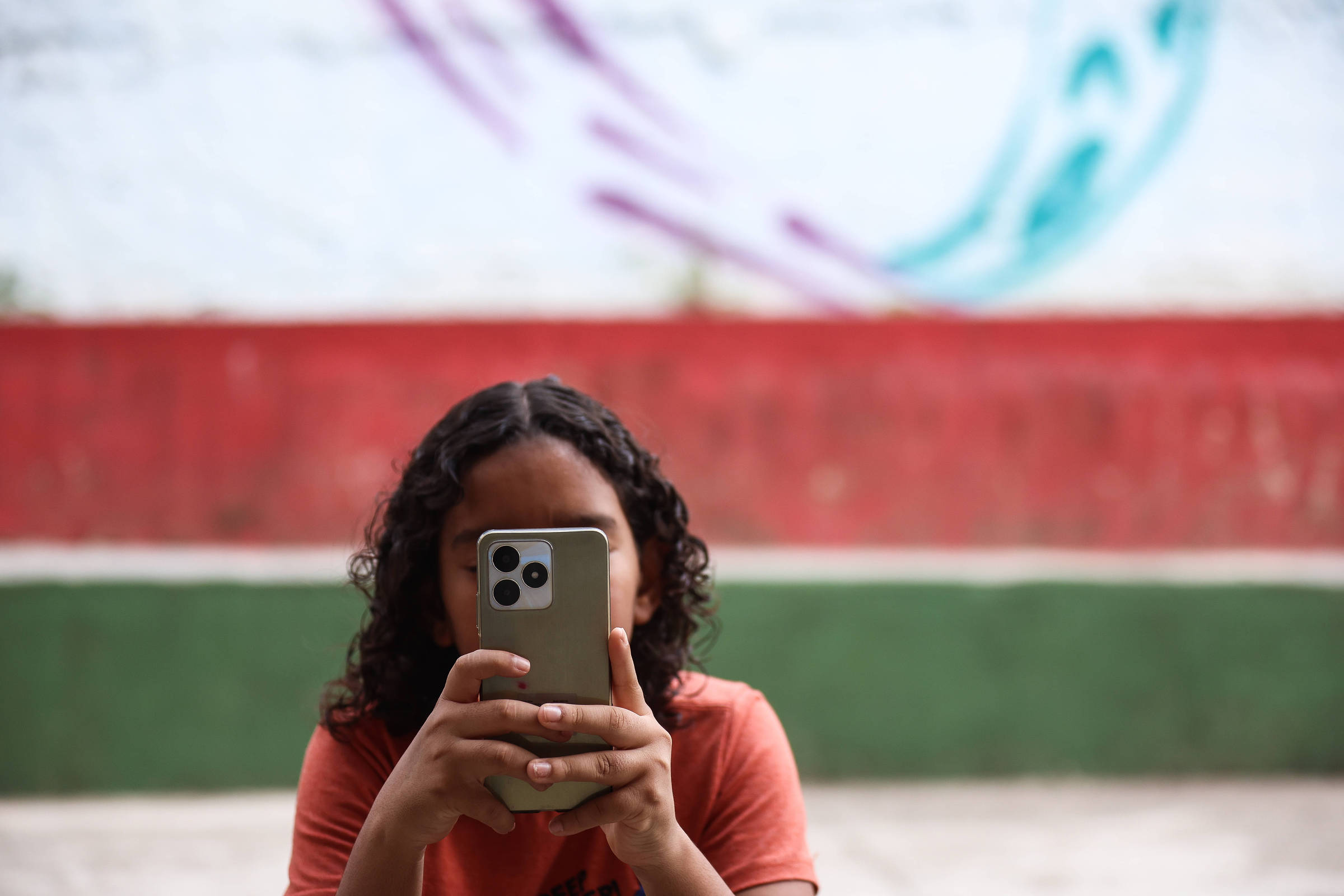Uma criança com cabelo cacheado e escuro está segurando um celular em frente ao rosto, olhando para a tela. Ao fundo, há uma parede com grafite colorido e bancos de concreto pintados em vermelho e verde.