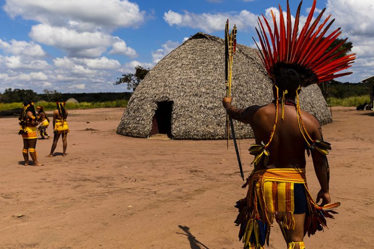 Um índio segura um arco com flecha com o braço erguido. Ao fundo, há uma oca e algumas mulheres conversando. O dia está ensolarado e o chão é de terra batida