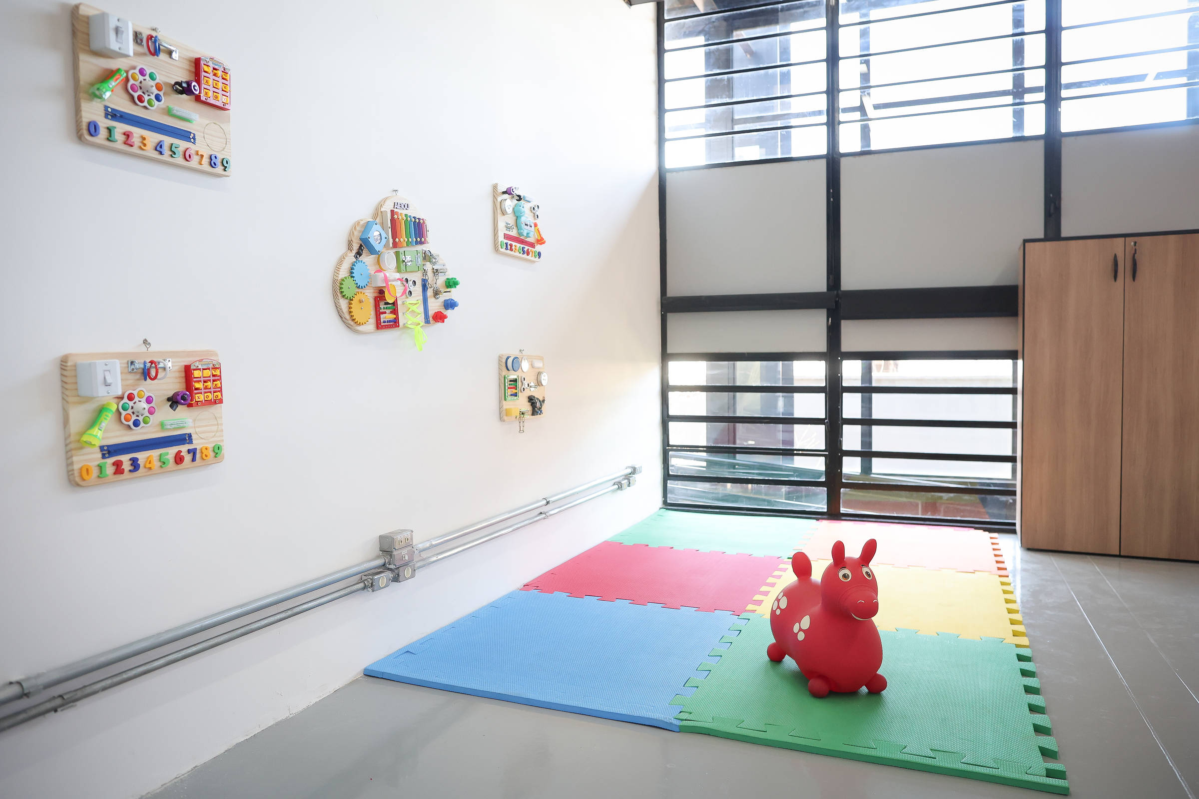A imagem mostra um ambiente de sala de brincadeiras com paredes claras e janelas grandes que permitem a entrada de luz natural. Na parede à esquerda, há painéis de atividades coloridos com botões e formas. No chão, há um tapete de espuma em cores variadas (azul, vermelho, verde e amarelo) e, em destaque, um brinquedo inflável em forma de cavalo vermelho. No fundo, há um armário de madeira claro.
