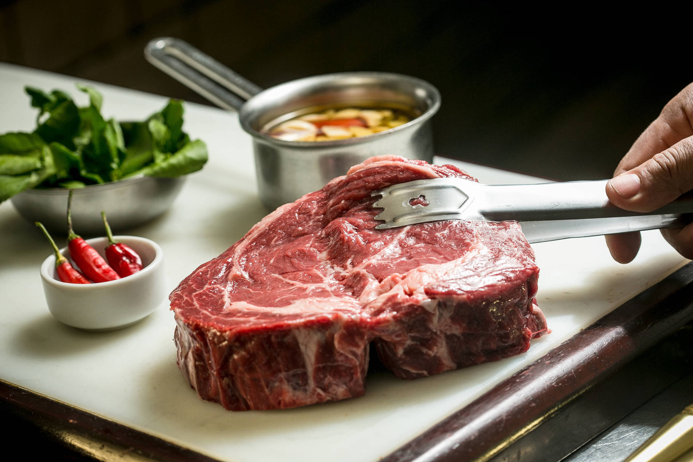 A imagem mostra um pedaço de carne crua, possivelmente um corte de costela, em uma tábua de corte. Ao lado, há um pequeno recipiente com molho e outro com folhas verdes. Também estão presentes algumas pimentas vermelhas em um recipiente separado. A mão de uma pessoa está segurando um garfo próximo à carne.