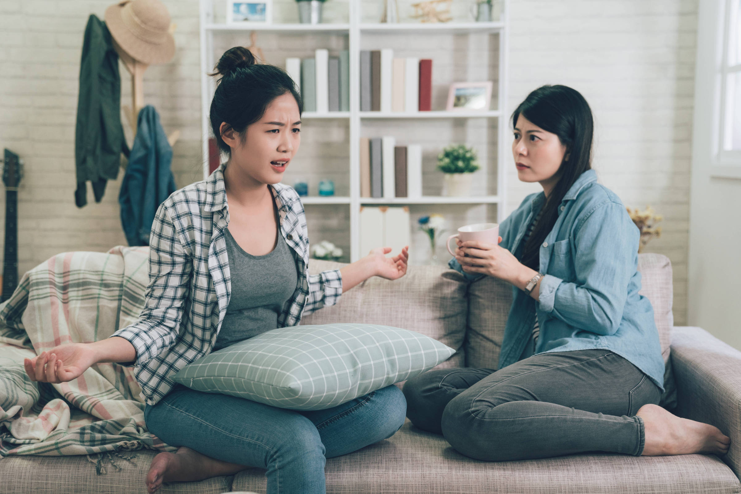 Duas mulheres estão sentadas em um sofá em uma sala de estar. A mulher à esquerda, com cabelo preso e usando uma camisa xadrez, parece estar expressando frustração ou descontentamento, gesticulando com as mãos. A mulher à direita, com cabelo solto e usando uma camisa jeans, observa atentamente, segurando uma xícara. O ambiente é acolhedor, com prateleiras ao fundo e uma manta sobre o sofá.