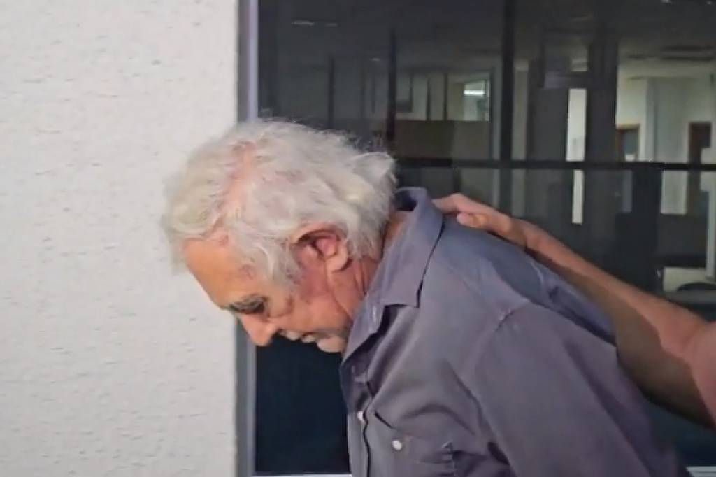 A imagem mostra um homem idoso com cabelo grisalho, caminhando com a cabeça baixa. Ele está usando uma camisa de botão cinza e é apoiado por uma mão que parece ser de outra pessoa. Ao fundo, há uma parede clara e janelas que indicam um ambiente interno.