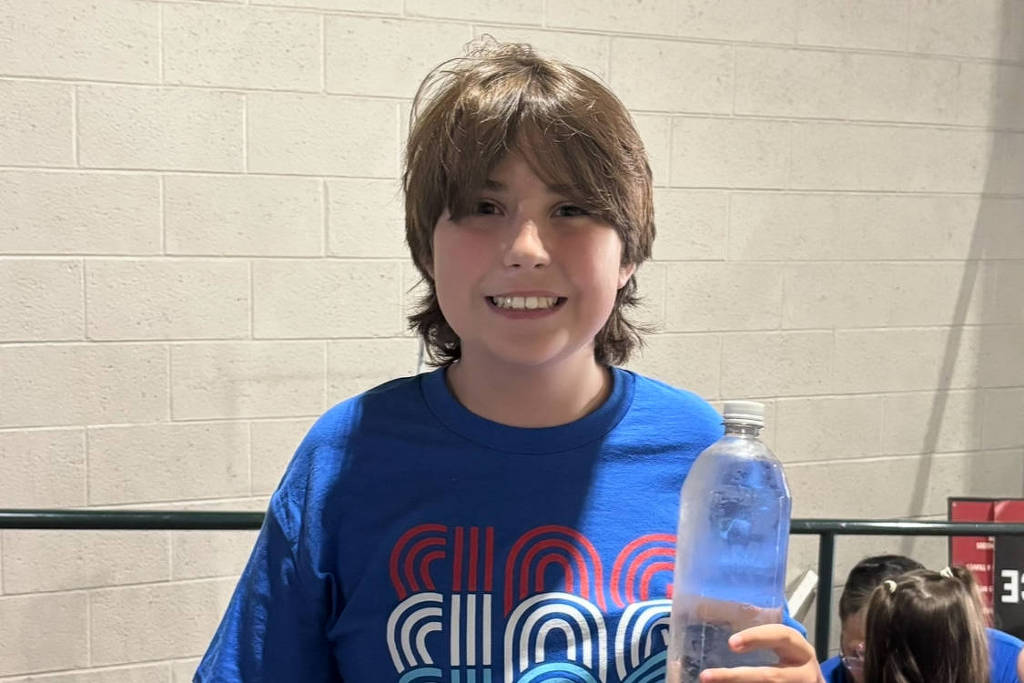 Um menino está sorrindo e segurando uma garrafa de água. Ele usa uma camiseta azul com a inscrição colorida