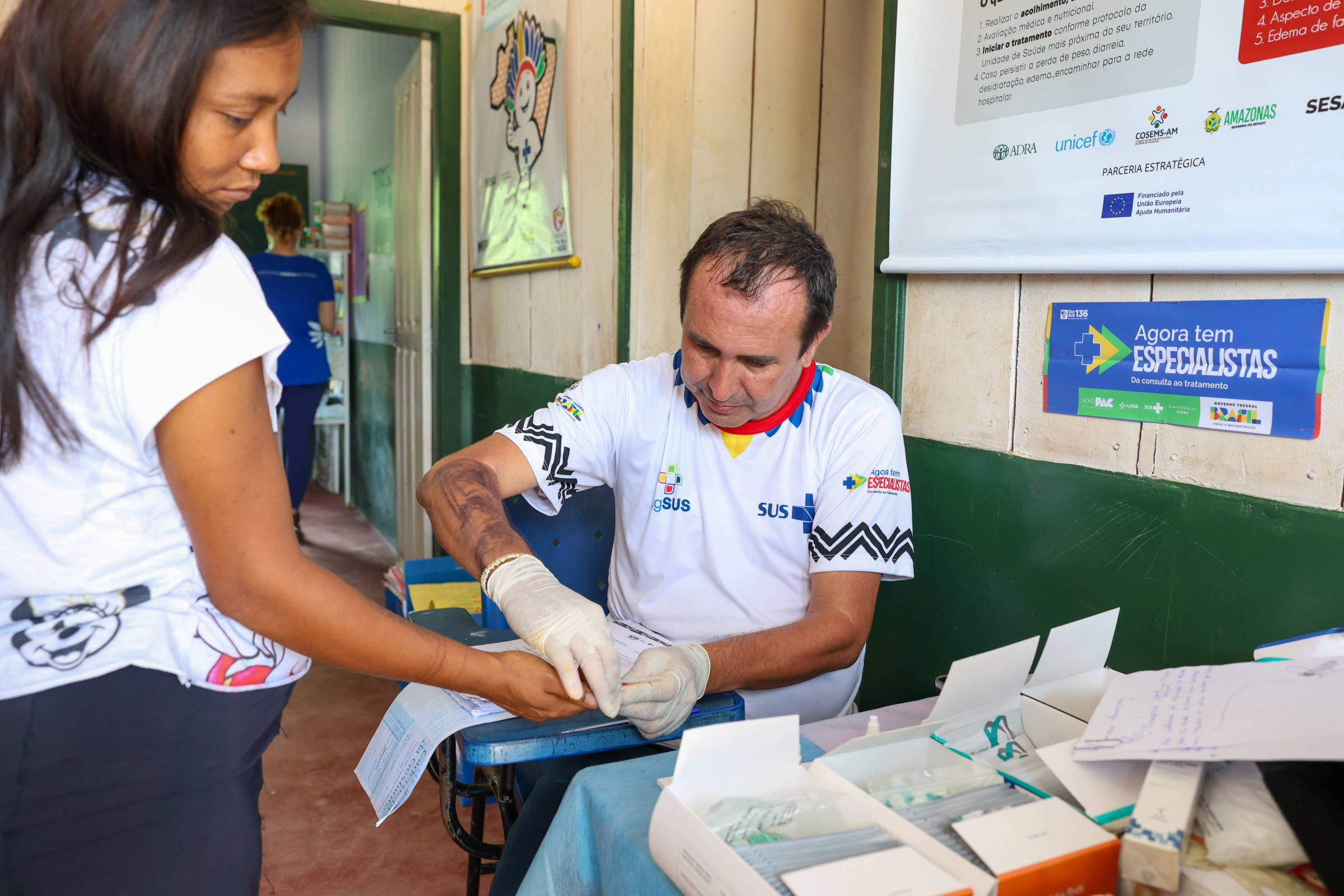 A imagem mostra um profissional de saúde, vestido com uma camisa do SUS, realizando um procedimento em uma paciente. A mulher, que está de pé, estende a mão para o profissional, que está sentado e usando luvas.