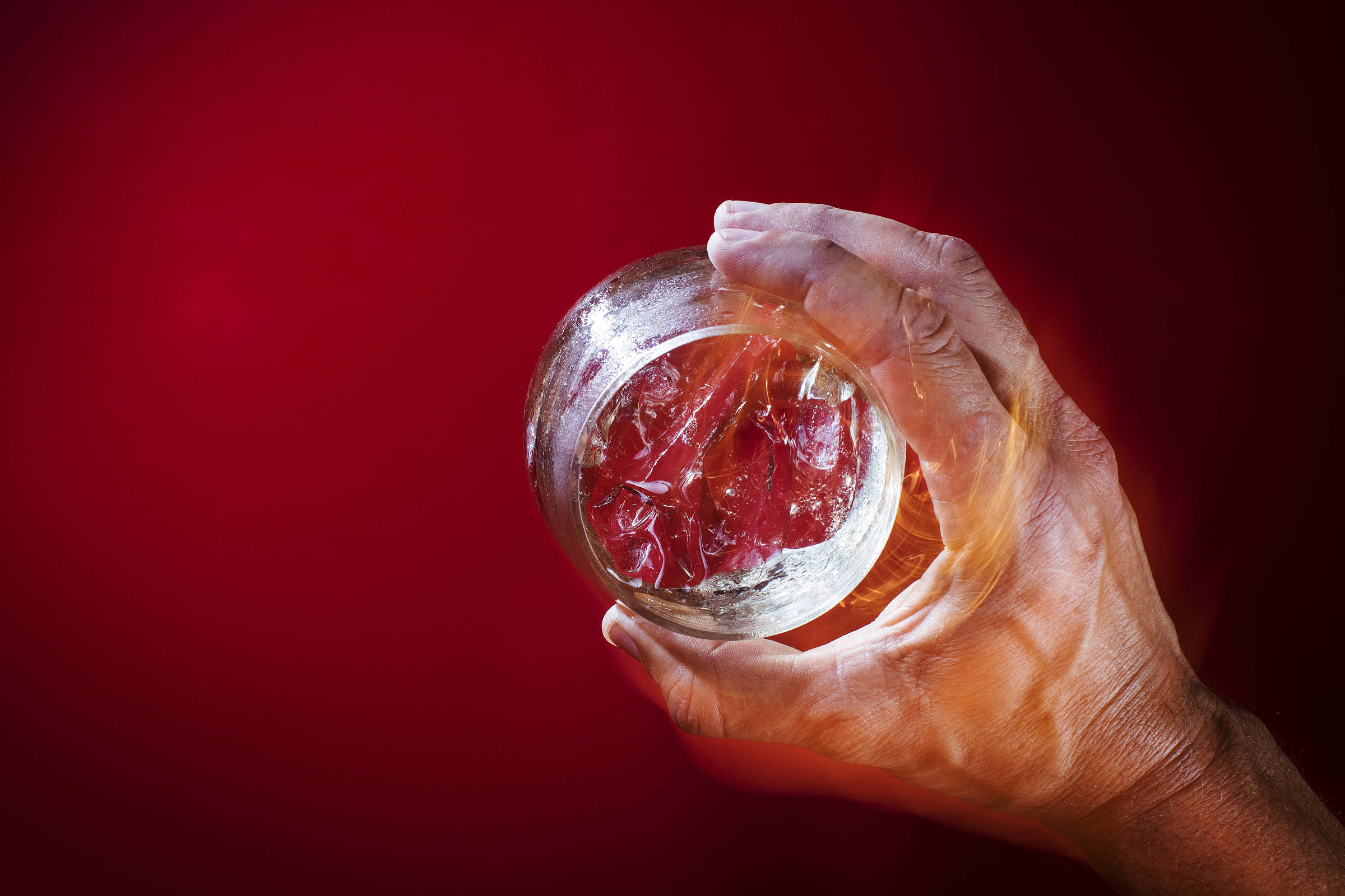 Mão direita segura um copo transparente visto de cima, contendo cubos de gelo. Fundo é vermelho sólido, sem outros elementos visíveis.
