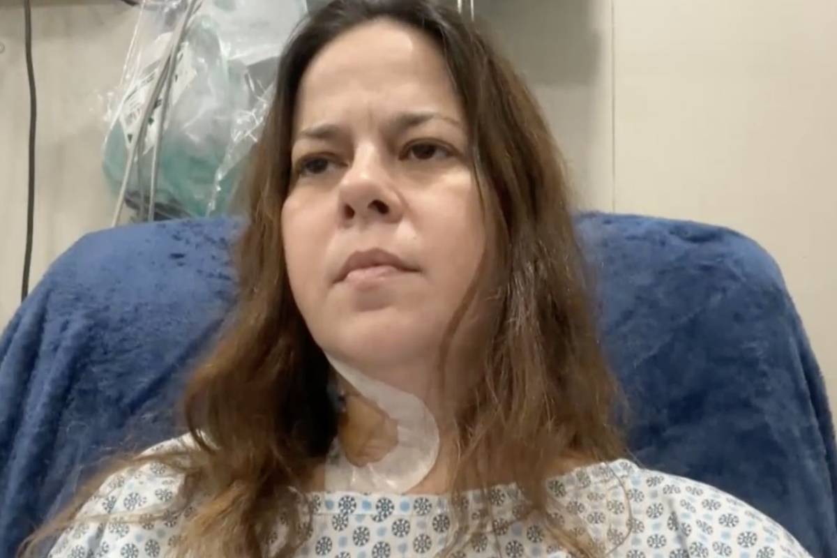 Mulher de cabelos longos castanhos sentada em cadeira com cobertor azul, vestindo roupa hospitalar branca com padrão azul. Curativo visível no pescoço. Equipamentos médicos ao fundo.