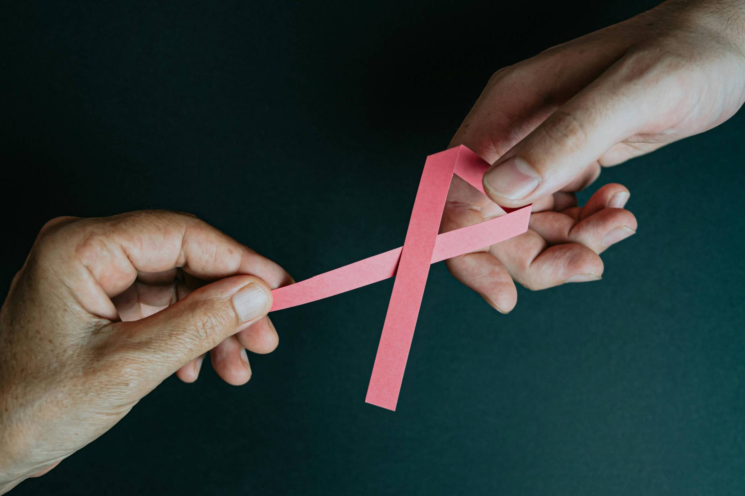 A imagem mostra duas mãos segurando uma fita rosa em forma de laço. A mão à esquerda é de pele mais escura e a mão à direita é de pele mais clara. O fundo é escuro, destacando o laço rosa.