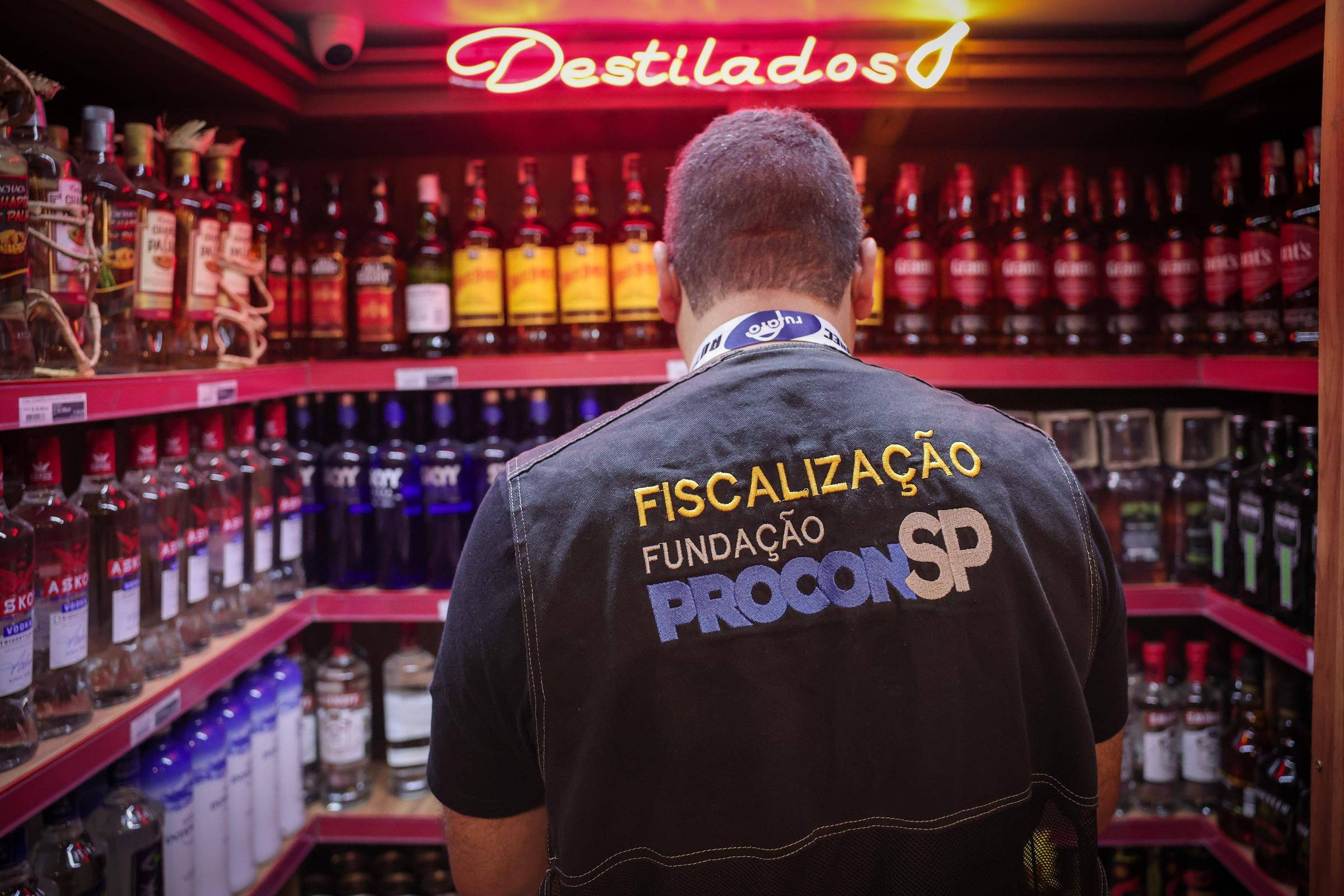 Agente da fiscalização do Procon-SP de costas, em frente a prateleiras cheias de garrafas de bebidas alcoólicas em loja. Acima, letreiro iluminado com a palavra