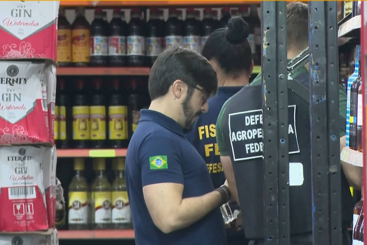 Dois agentes da fiscalização agropecuária federais inspecionam prateleiras de bebidas alcoólicas em supermercado. Um deles veste camisa azul com bandeira do Brasil na manga, o outro camisa preta com identificação
