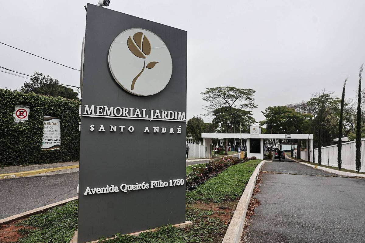 Placa cinza escuro com logotipo de flor branca e dourada indica Memorial Jardim Santo André na Avenida Queirós Filho, 1750. Ao fundo, portaria branca com guarita e veículos na entrada, cercada por vegetação e árvores sob céu nublado.
