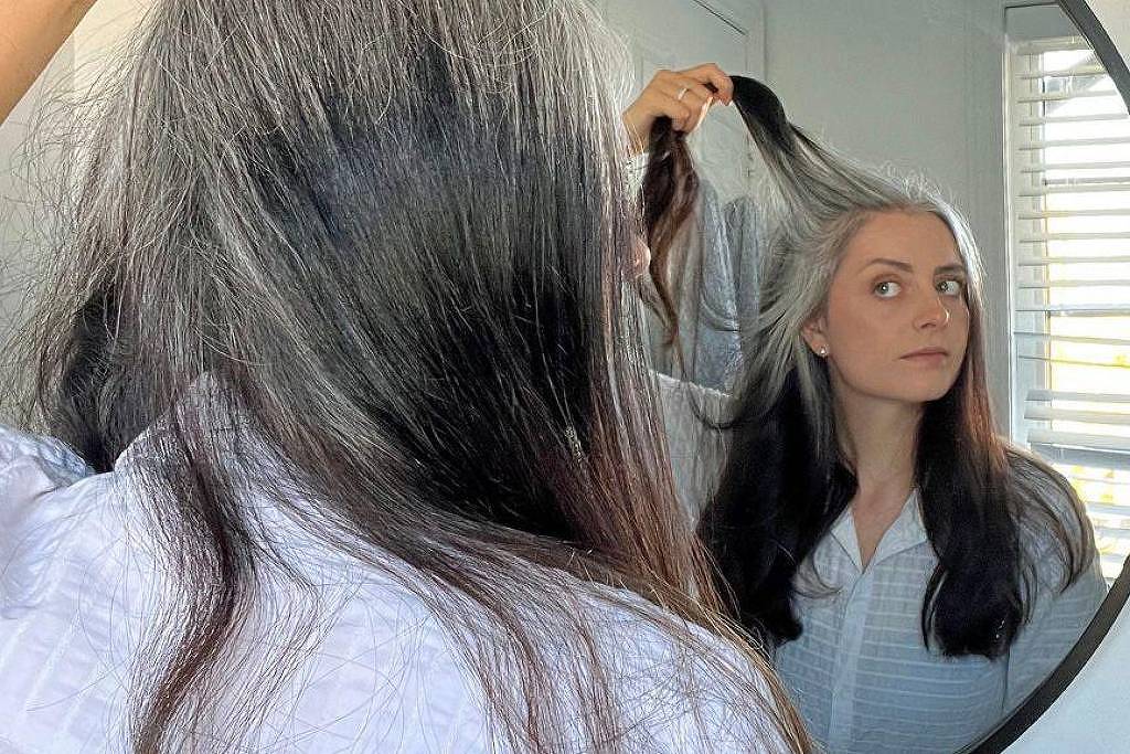 Mulher com cabelo longo e bicolor, metade preto e metade grisalho, segura mecha grisalha enquanto se olha em espelho redondo. Ela veste camisa clara e está em ambiente com janela de persiana ao fundo.