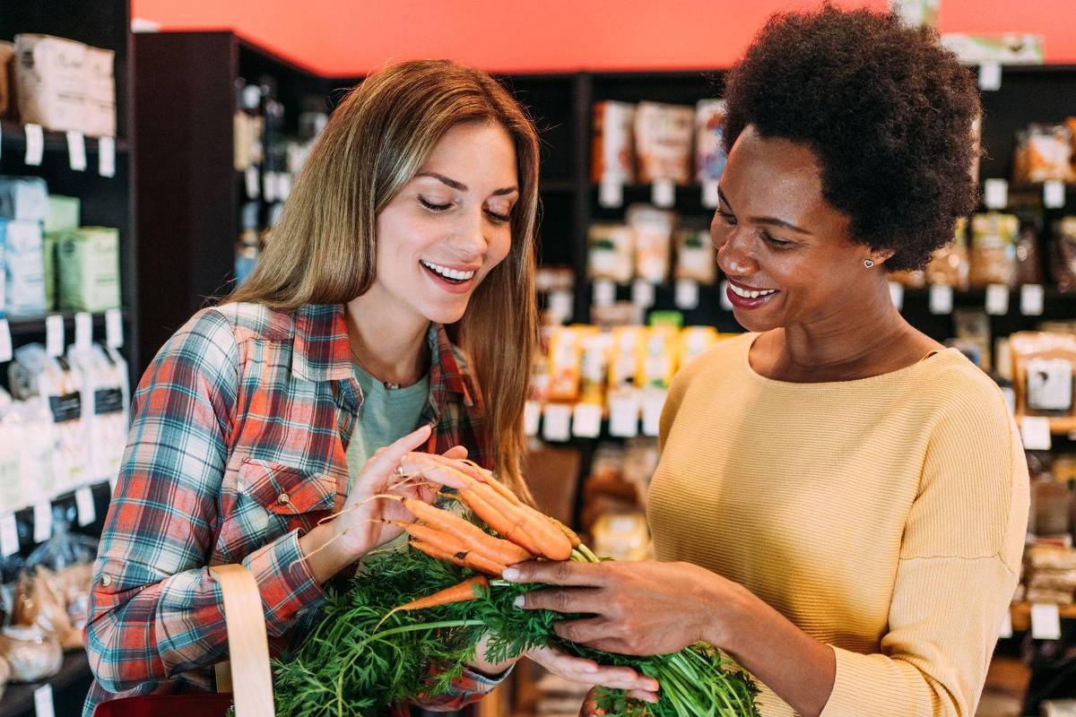 Duas mulheres em mercado de alimentos naturais seguram cenouras frescas. Uma veste camisa xadrez e a outra blusa amarela, sorrindo enquanto examinam o produto.