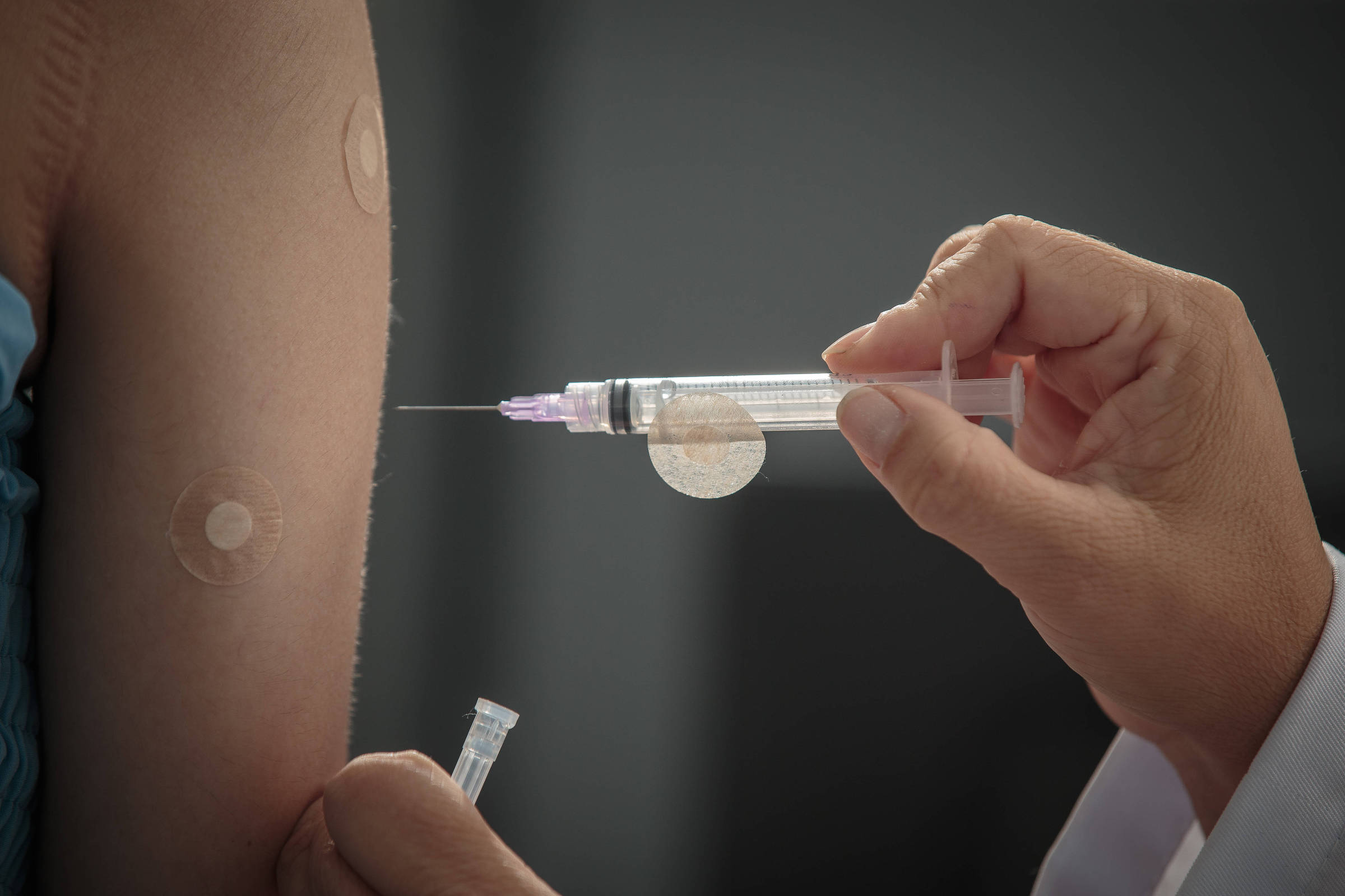 A imagem mostra uma mão segurando uma seringa com uma agulha, prestes a aplicar uma vacina em um braço humano. O braço está exposto e possui adesivos circulares, possivelmente de vacinas anteriores. O fundo é desfocado, focando na ação da vacinação.