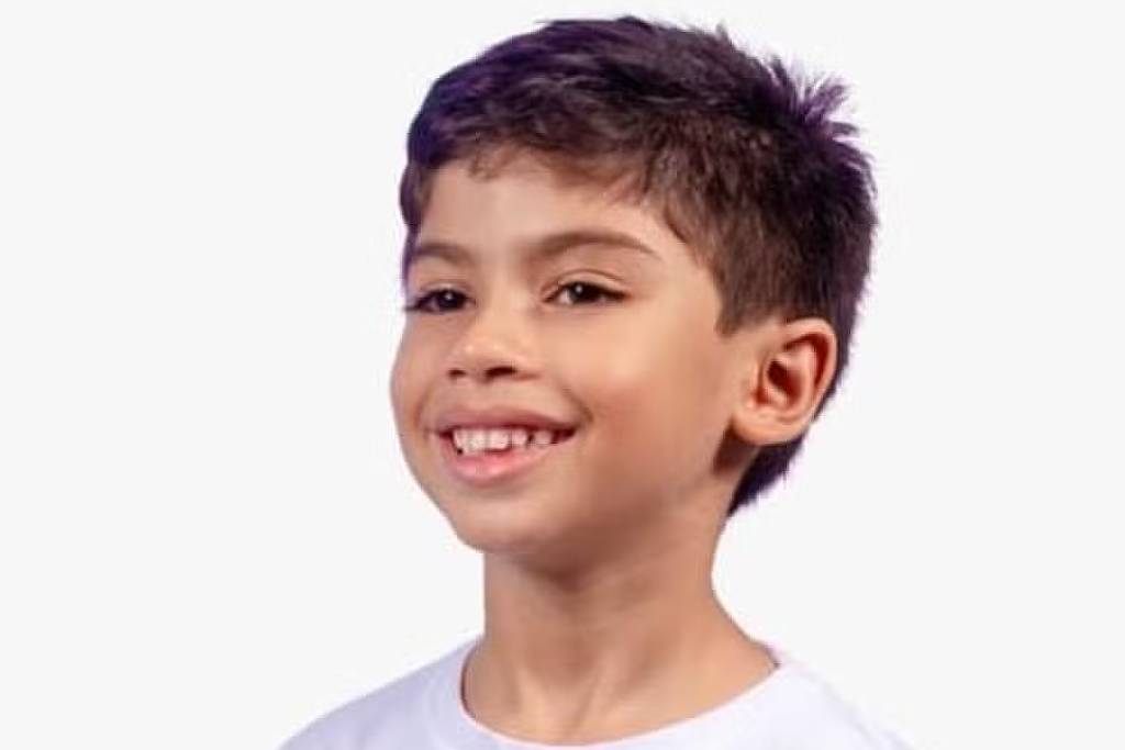 Menino de cabelo curto e escuro sorri levemente, vestindo camiseta branca com estampa colorida que mostra uma cena urbana com figuras e prédios. Fundo é branco e uniforme.