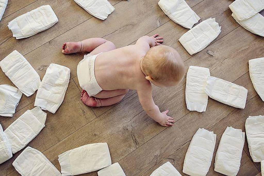 Bebê de costas, usando apenas fralda, sentado no chão de madeira cercado por várias fraldas descartáveis espalhadas ao redor.