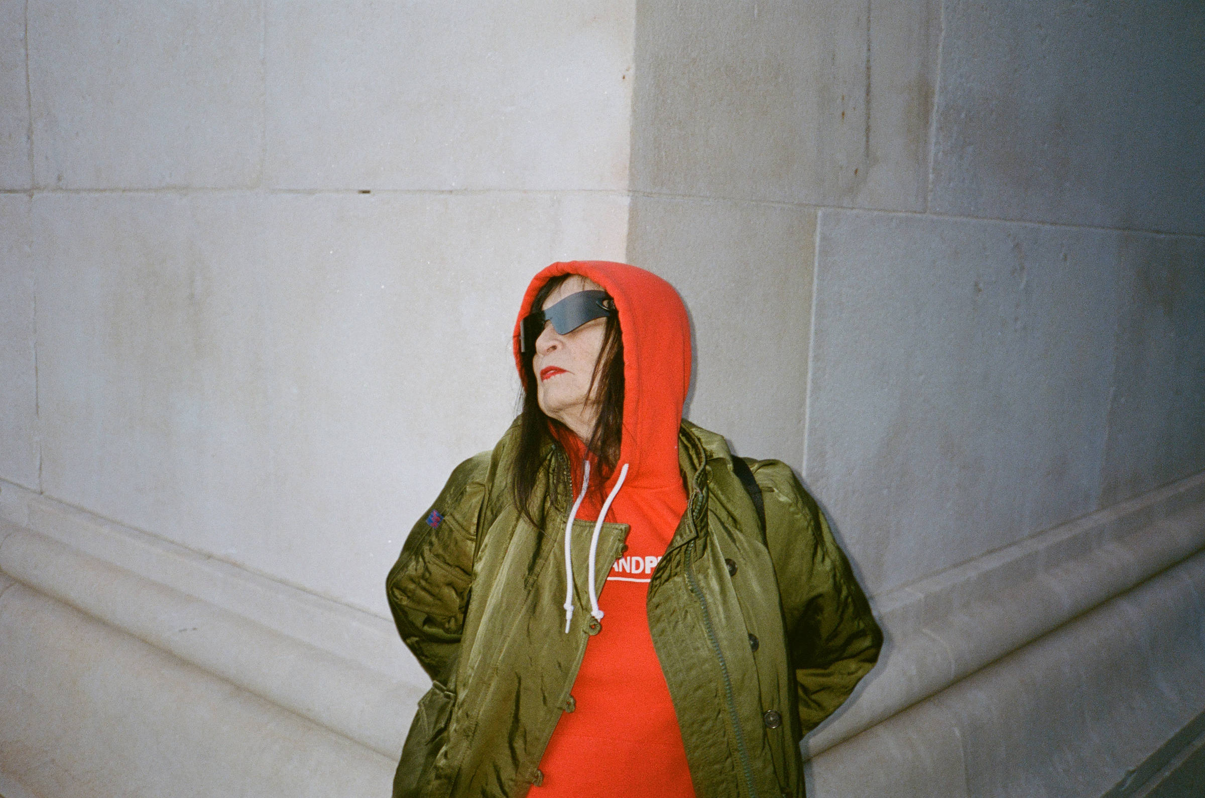Pessoa com capuz vermelho e jaqueta verde escura encostada em canto de parede de pedra clara. Usa óculos escuros e olha para o lado direito da imagem.