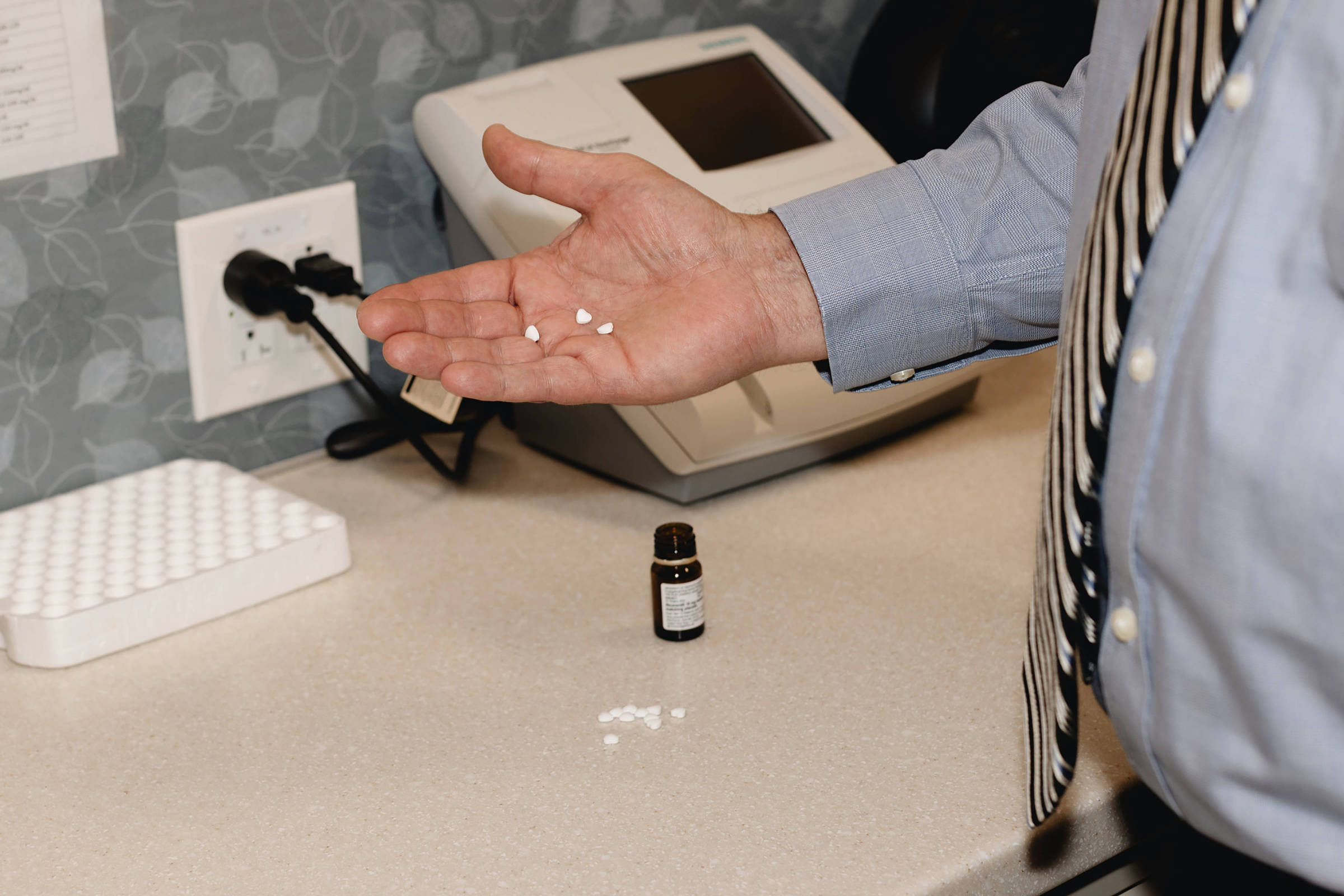 Pessoa com camisa azul e gravata segura comprimidos brancos na palma da mão sobre bancada com frasco pequeno e equipamento médico ao fundo.