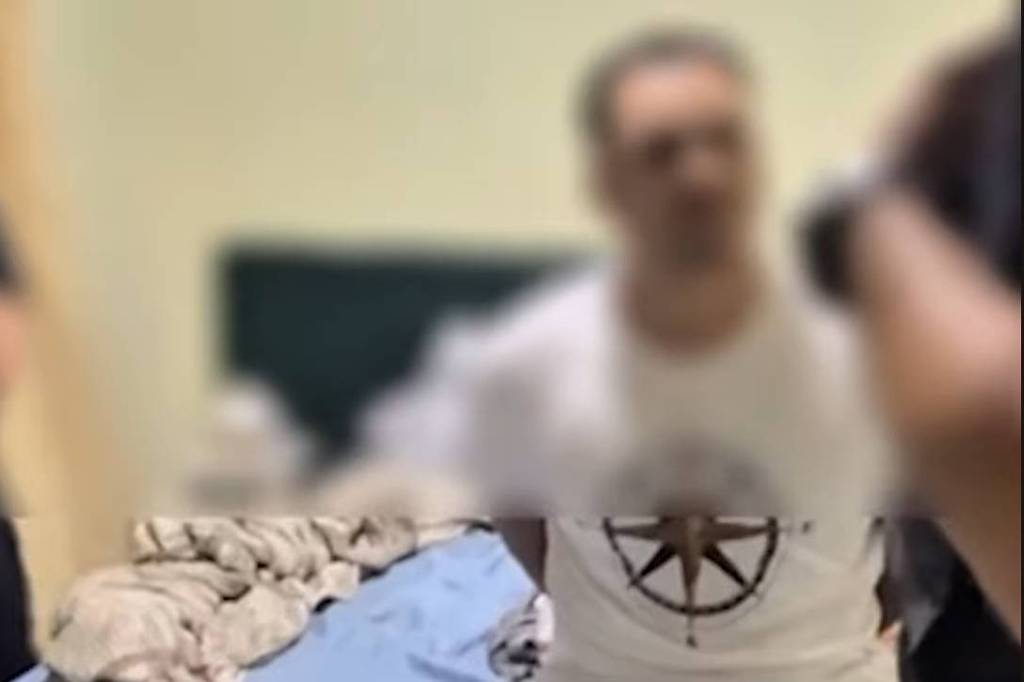 Homem de camiseta branca e bermuda clara sentado na beira de uma cama com lençol azul e cobertor amontoado. Ambiente interno com parede clara e pouca decoração visível.