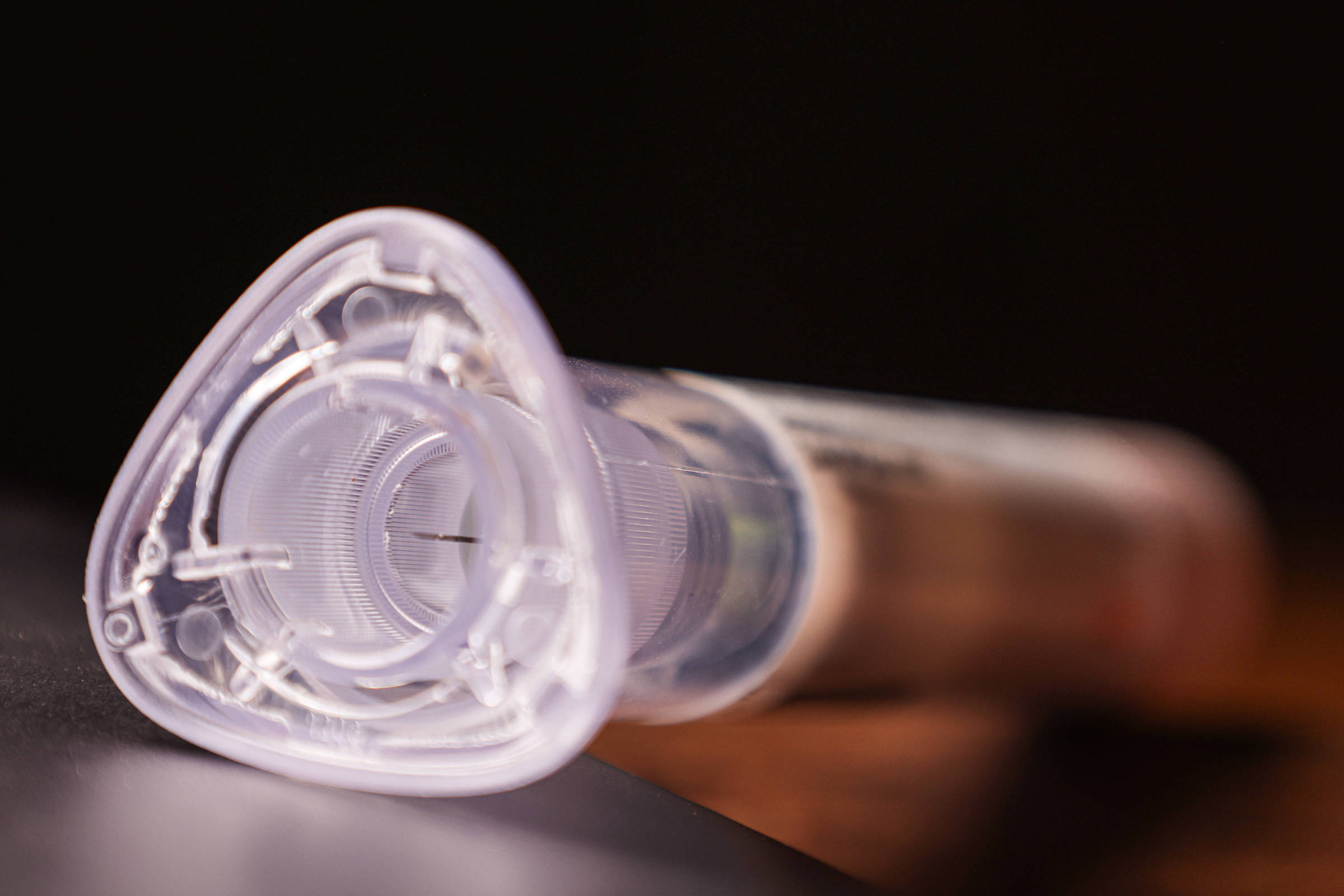 A imagem mostra uma seringa em close, com foco na parte superior que tem uma forma triangular. A seringa é transparente e apresenta detalhes internos visíveis, como um êmbolo e uma agulha. O fundo é desfocado, sugerindo um ambiente escuro.