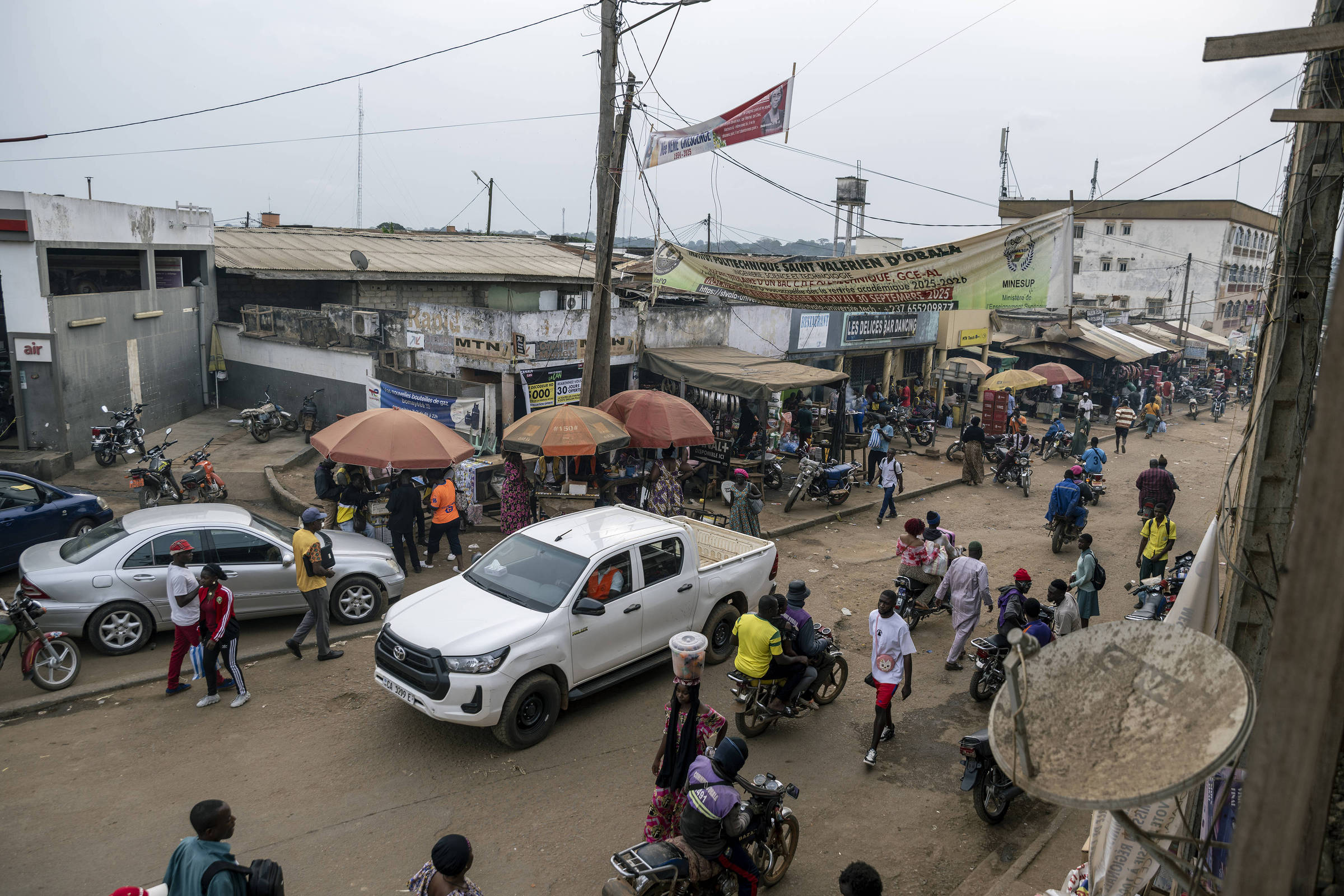 Rua movimentada com pedestres, motociclistas e carros em área comercial. Barracas com guarda-sóis e lojas ao fundo, sob céu nublado.